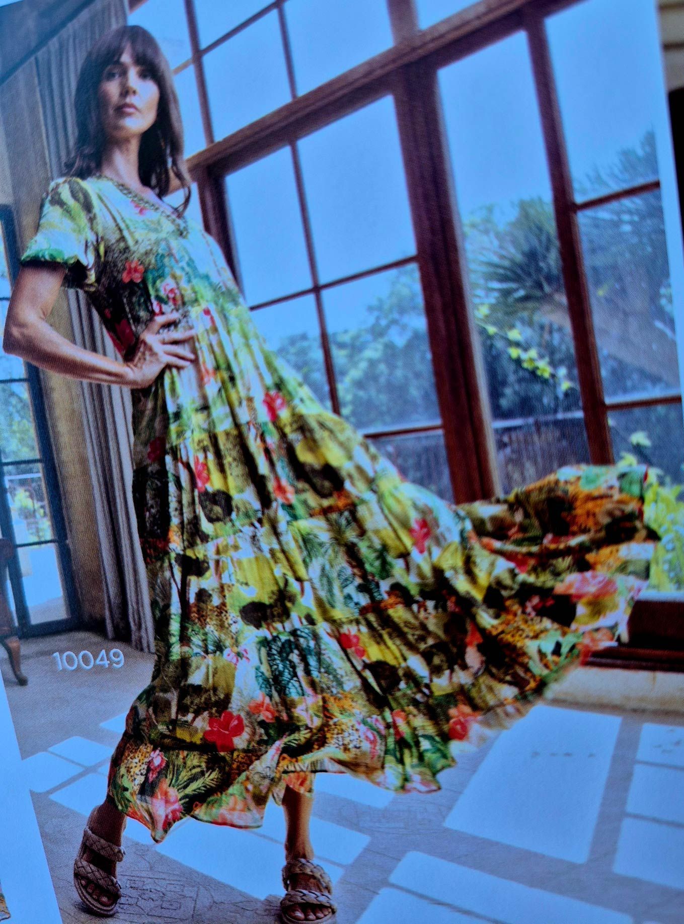 Woman in a flowing, colorful dress poses by a large window.