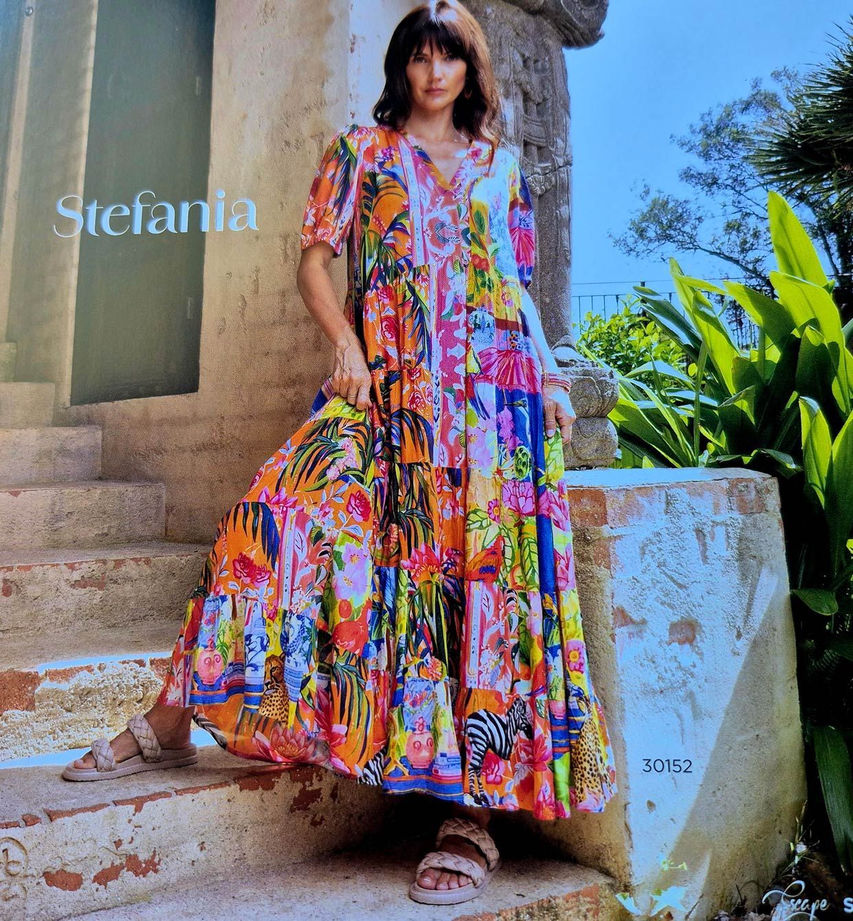 Woman in a colorful floral maxi dress stands on steps. Stone columns and plants in the background.