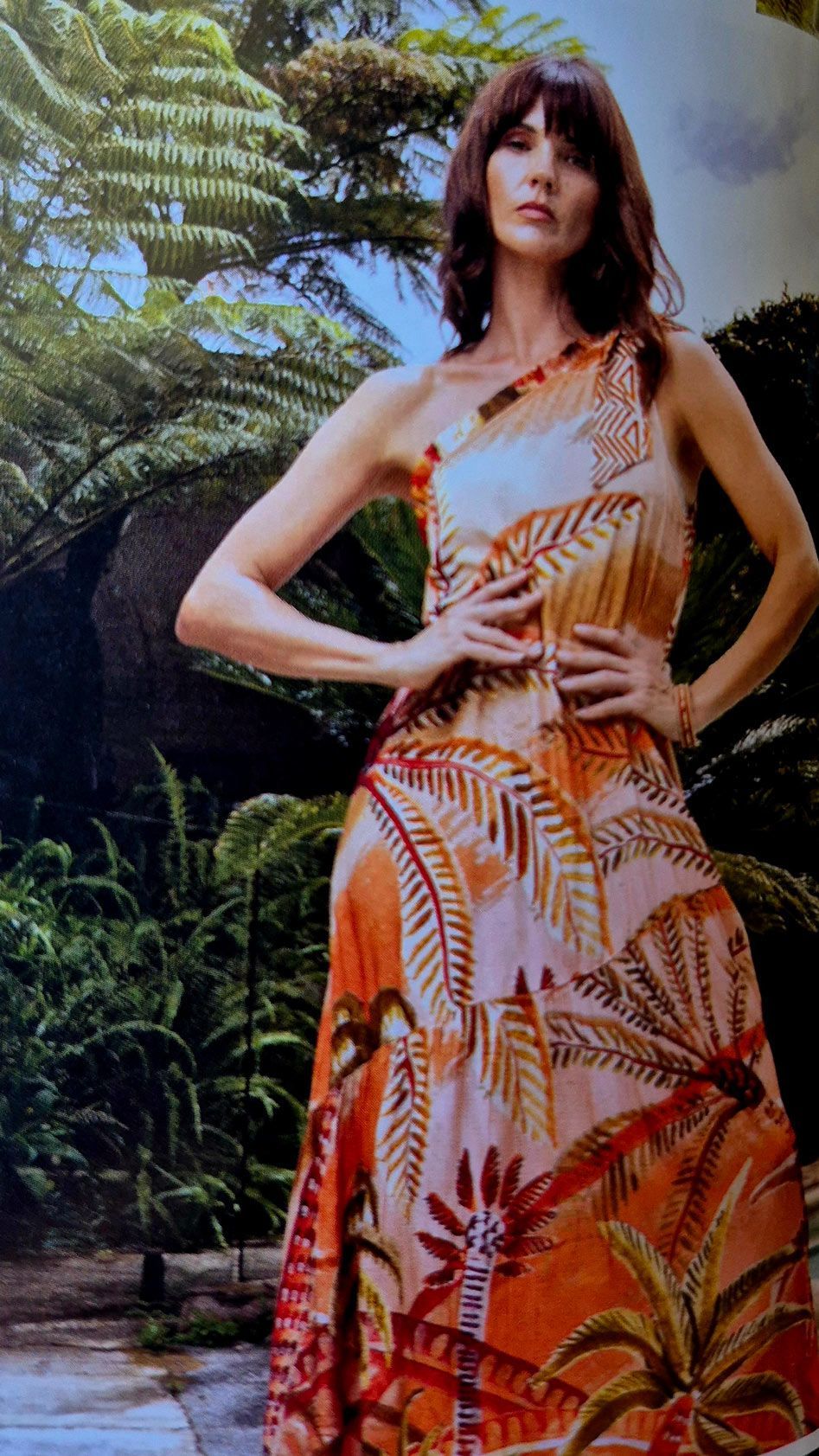Woman in a tropical print, one-shoulder dress, posing with hands on hips in an outdoor garden setting.