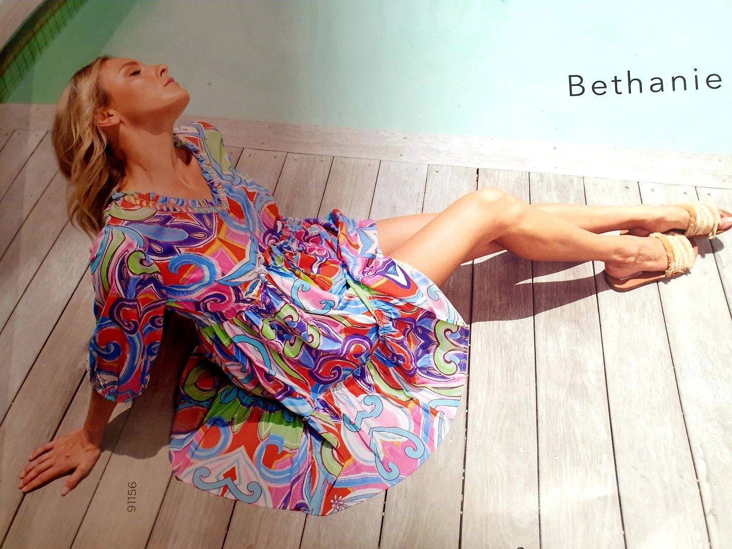 Woman in colorful dress, reclining outdoors on wooden planks, eyes closed, basking in sun.