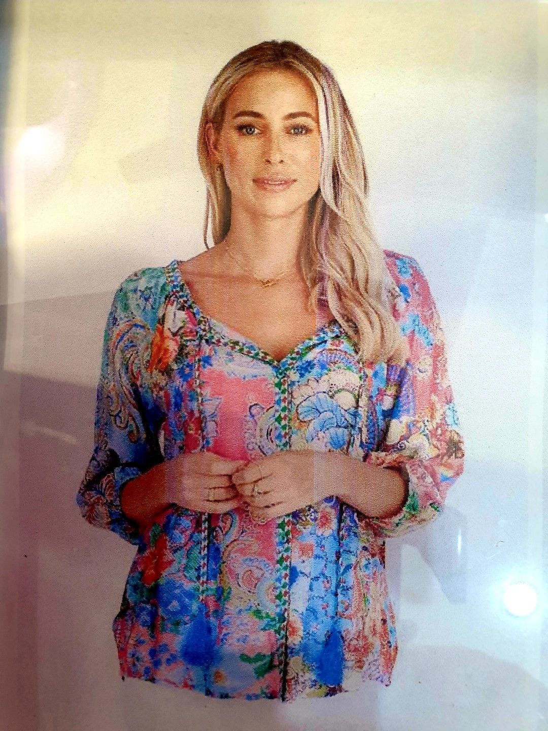 Blonde woman in floral top smiles, hands clasped, against a white background.