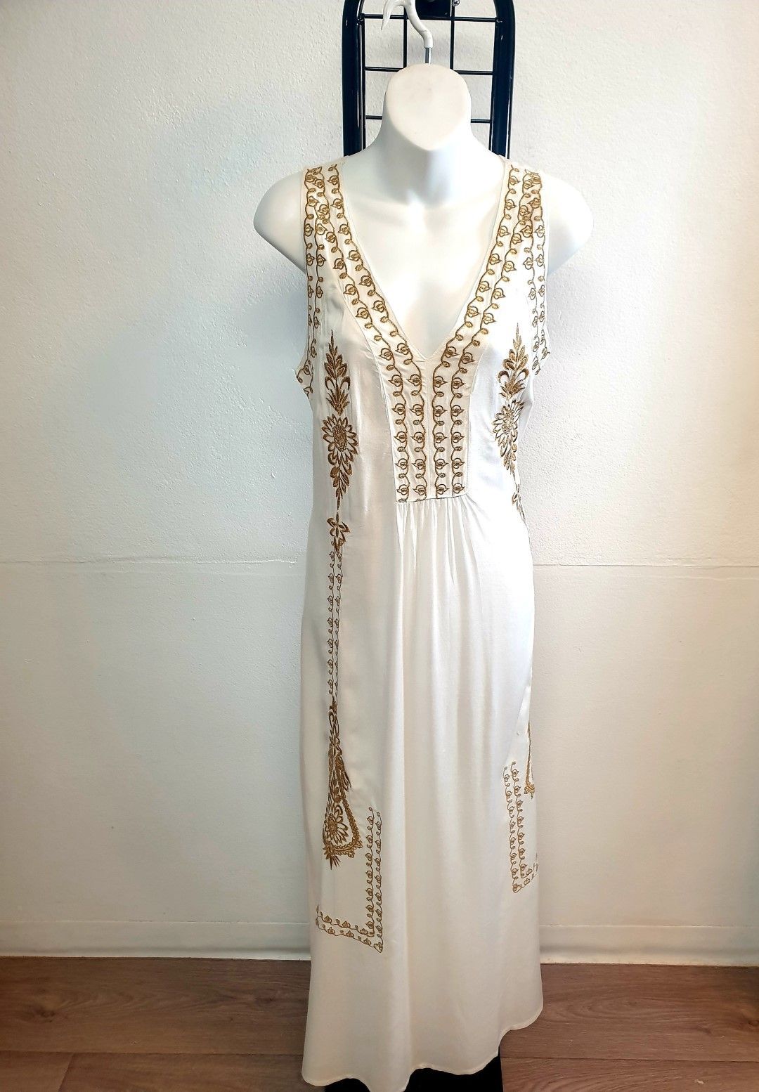 White sleeveless dress on a mannequin with gold embroidery, set against a white wall and wooden floor.