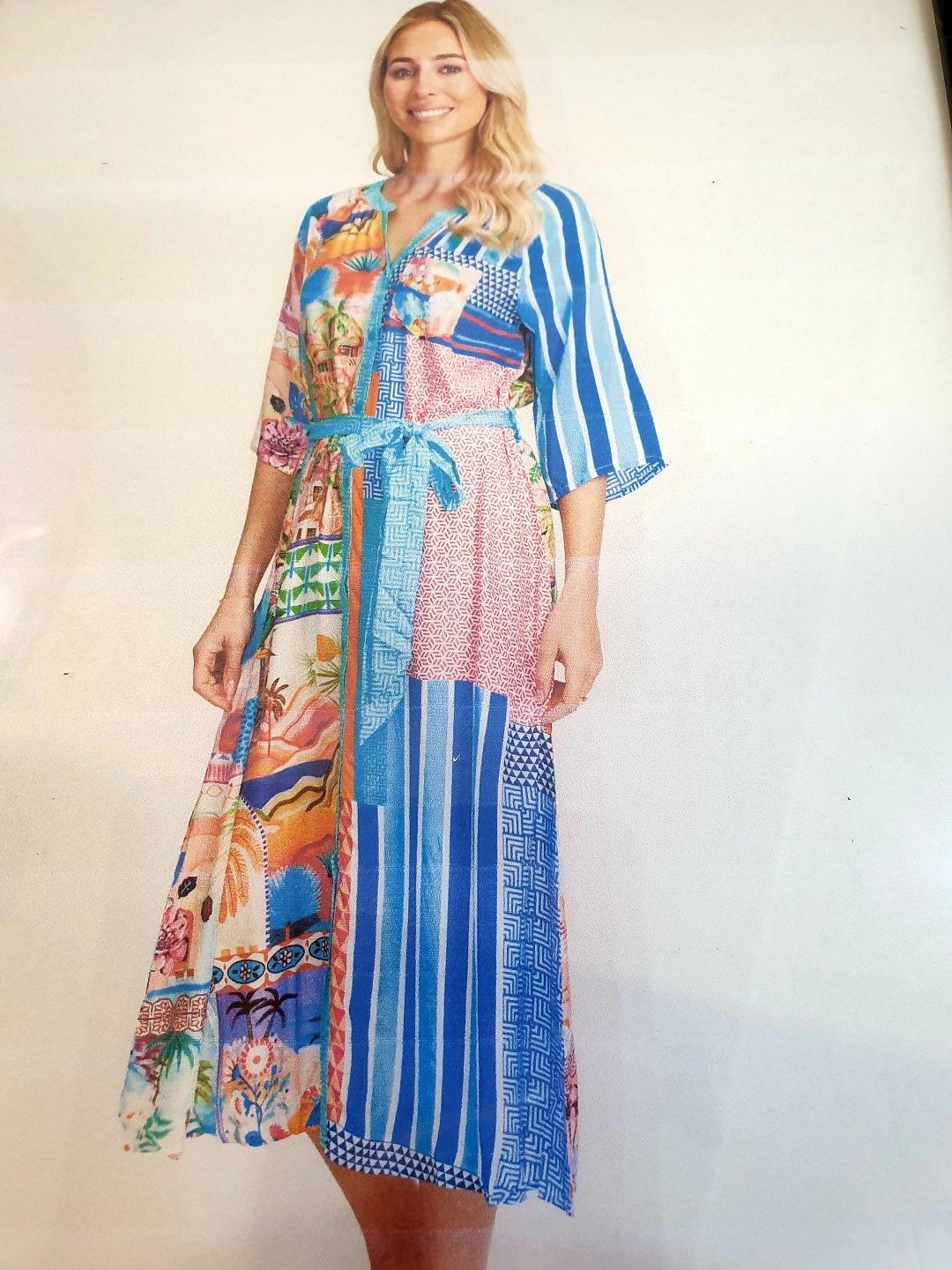 Woman in colorful patchwork dress smiling, standing against white wall.