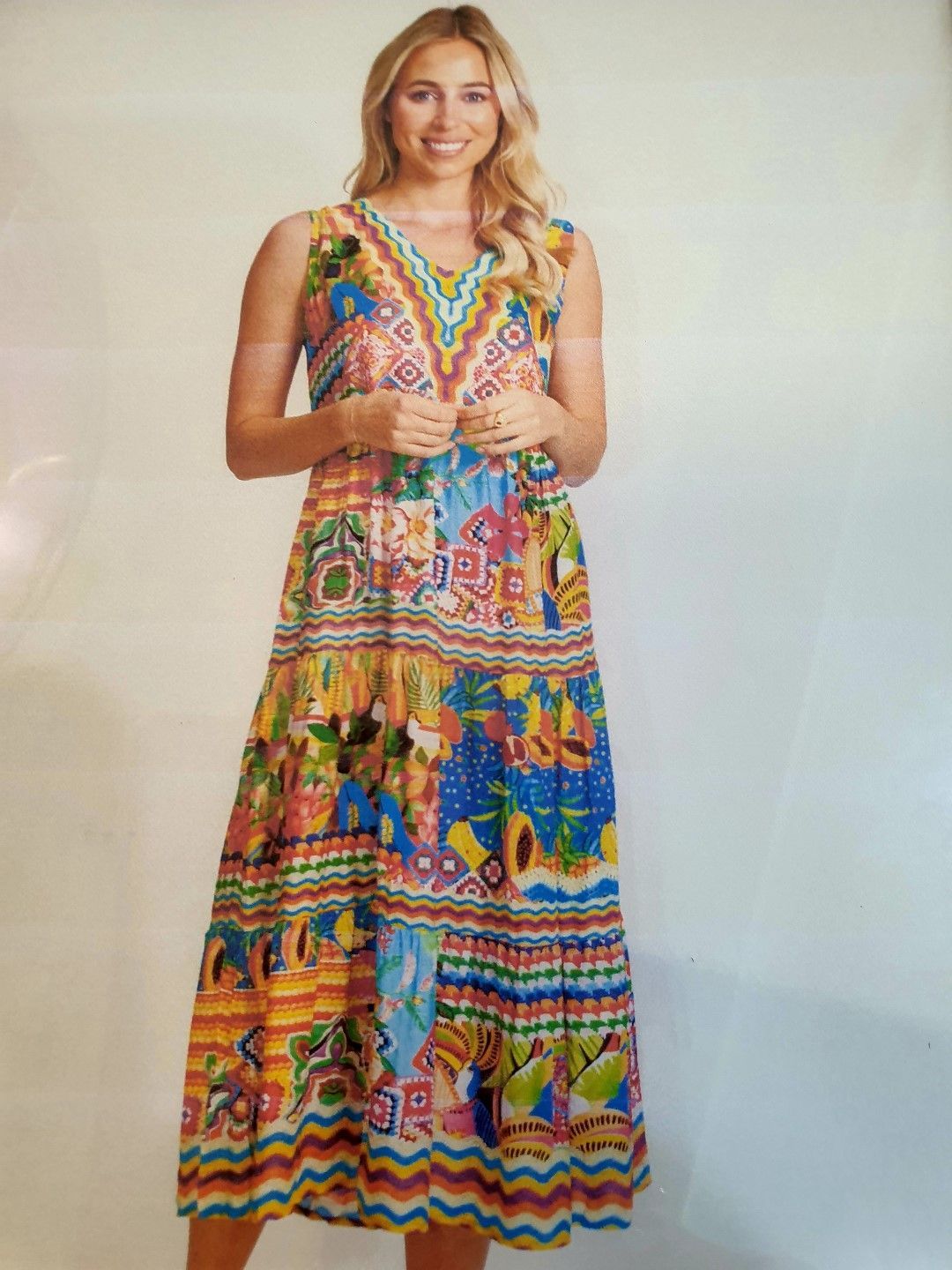Woman in a colorful sleeveless maxi dress smiles. She stands indoors against a light background.