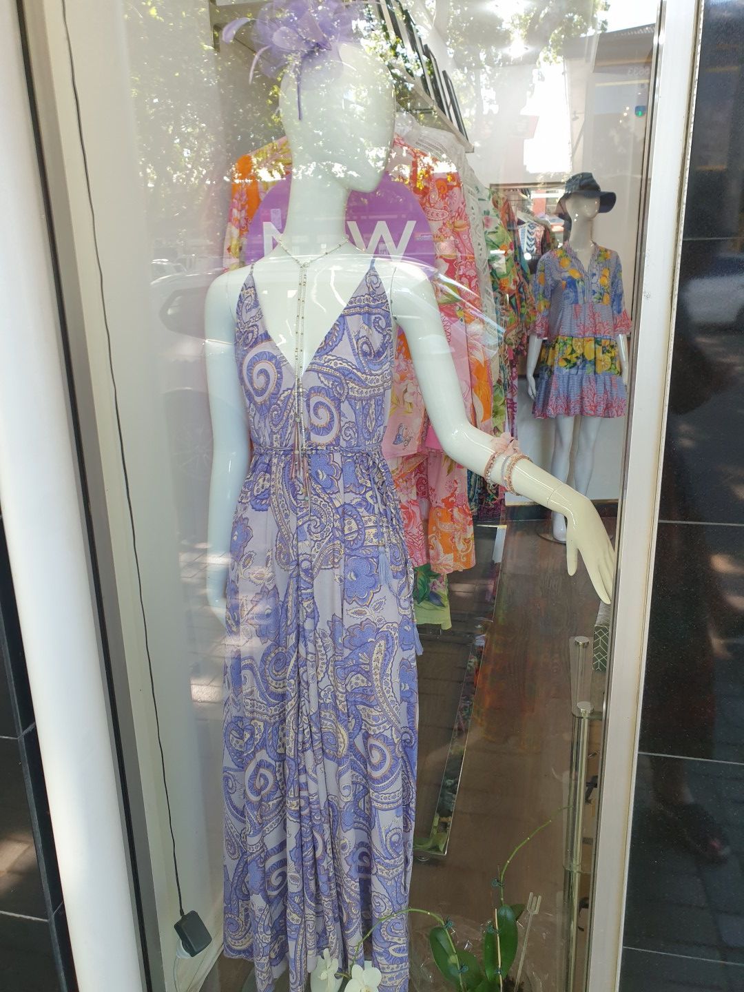 Mannequin in a lavender patterned dress and hat in a store window; other clothing visible in the background.