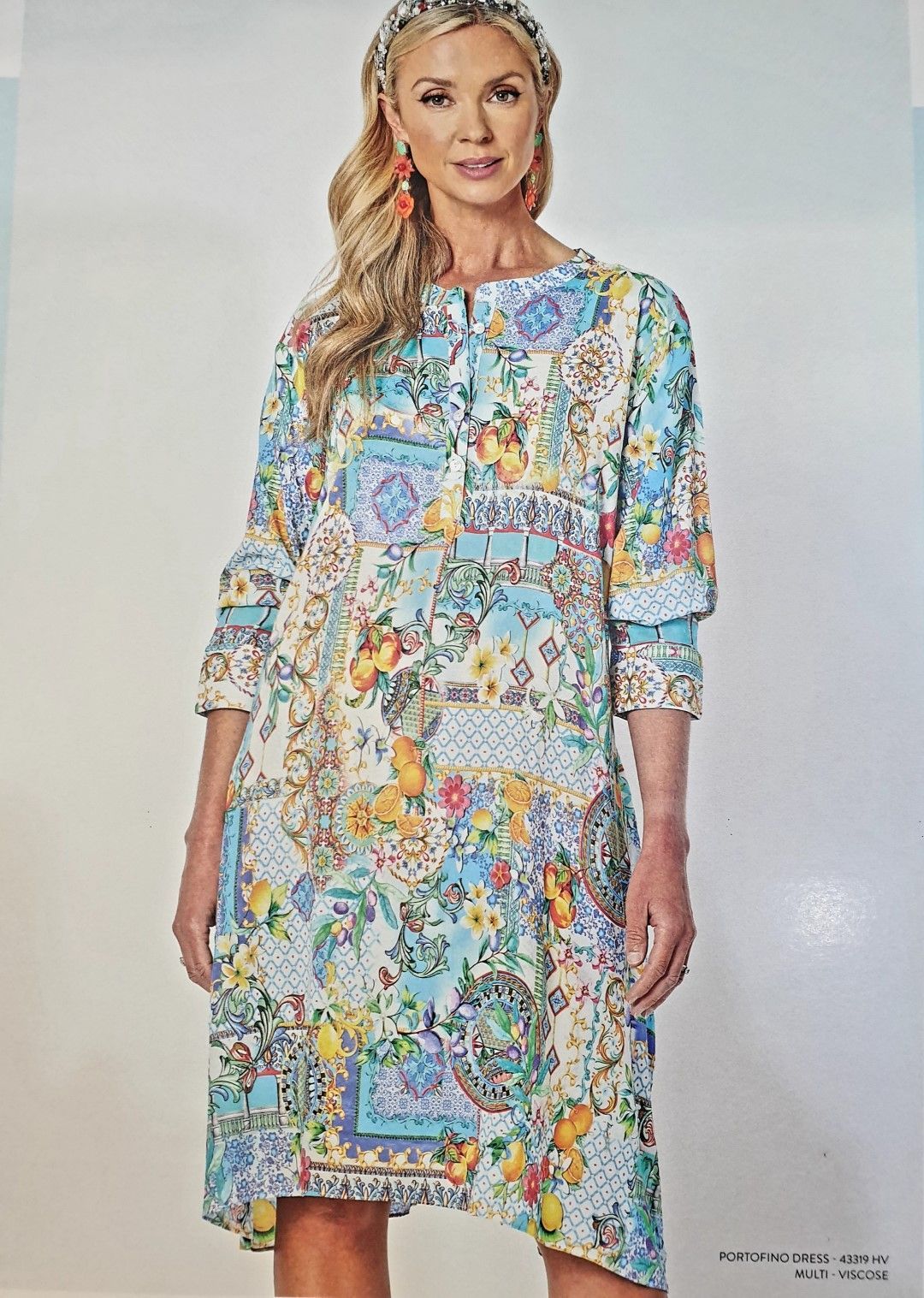 Woman wearing a colorful, patterned dress with a headband and earrings, standing in a light-filled setting.