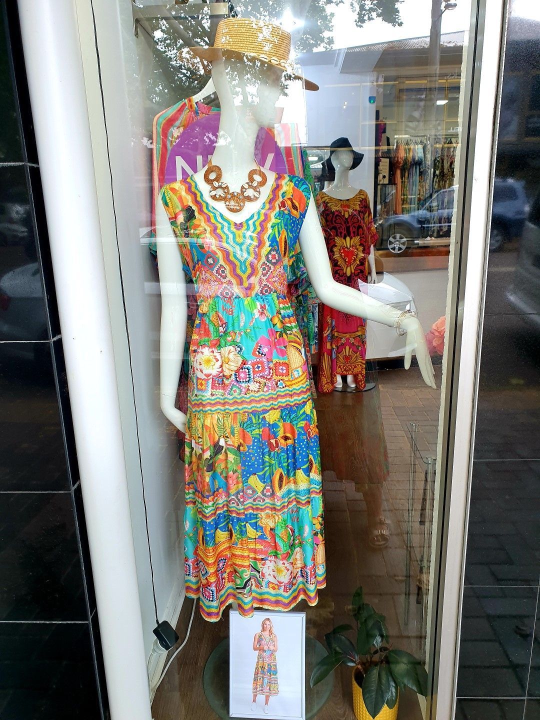 Mannequin wearing a colorful floral dress, necklace, and straw hat in a storefront window.