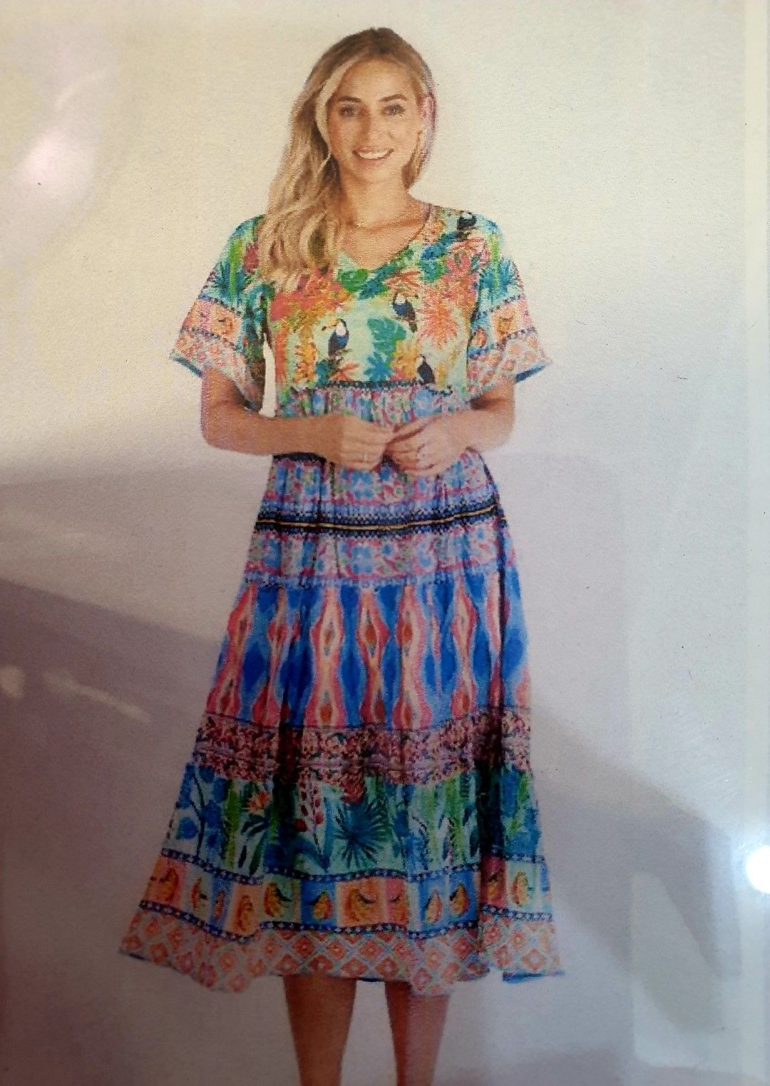 Woman wearing a colorful, floral dress; standing indoors with hands clasped and smiling.
