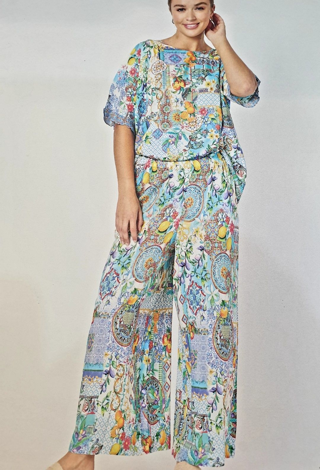 Woman in a patterned blue and white jumpsuit, smiling, standing with her hand on her head.