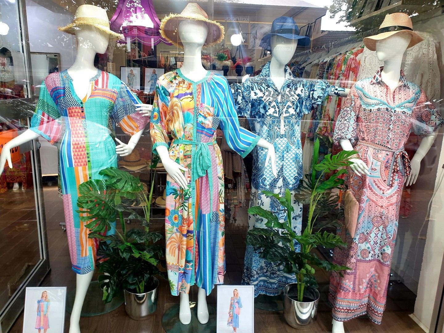 Four mannequins in dresses and hats in a shop window. Colorful dresses, green plants, natural light.