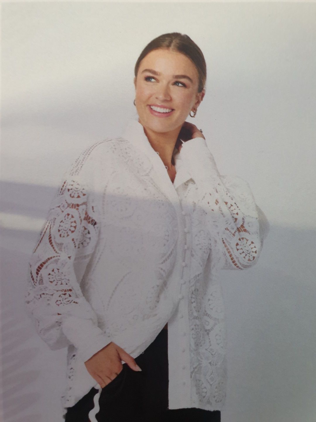 Woman in white eyelet shirt, smiling, hand in pocket, in a studio.