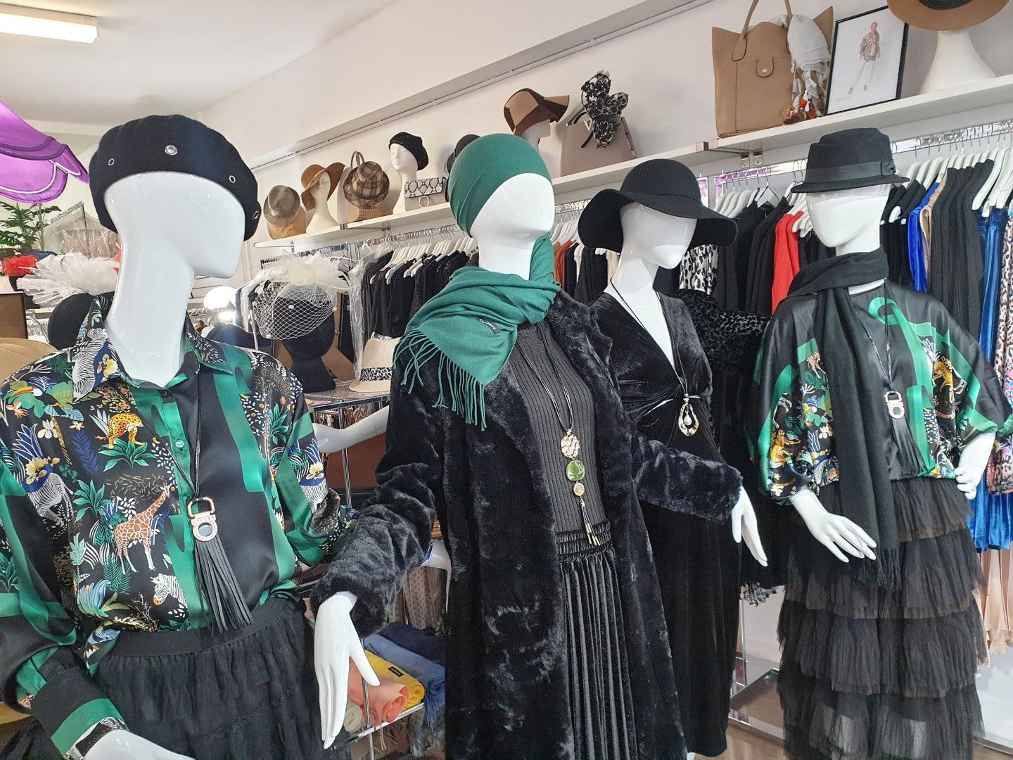 Clothing store display with mannequins wearing hats, coats, and skirts. Black, green, and floral prints are featured.
