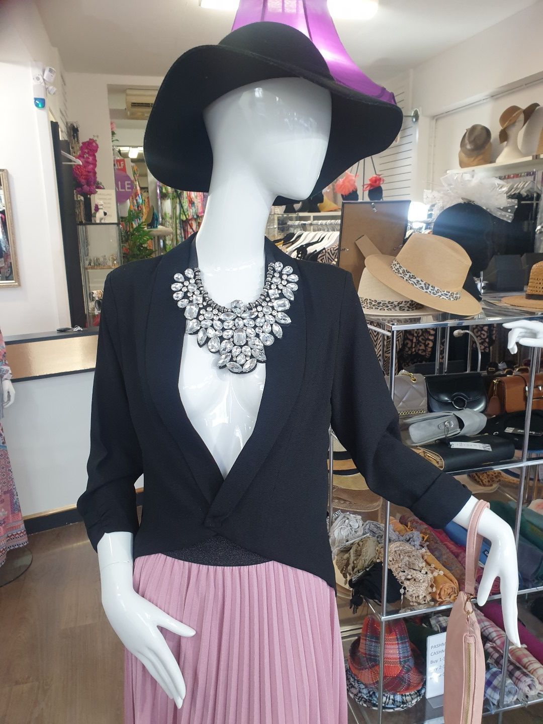 Mannequin wearing a black hat, sweater, statement necklace, and pink skirt; inside a boutique.