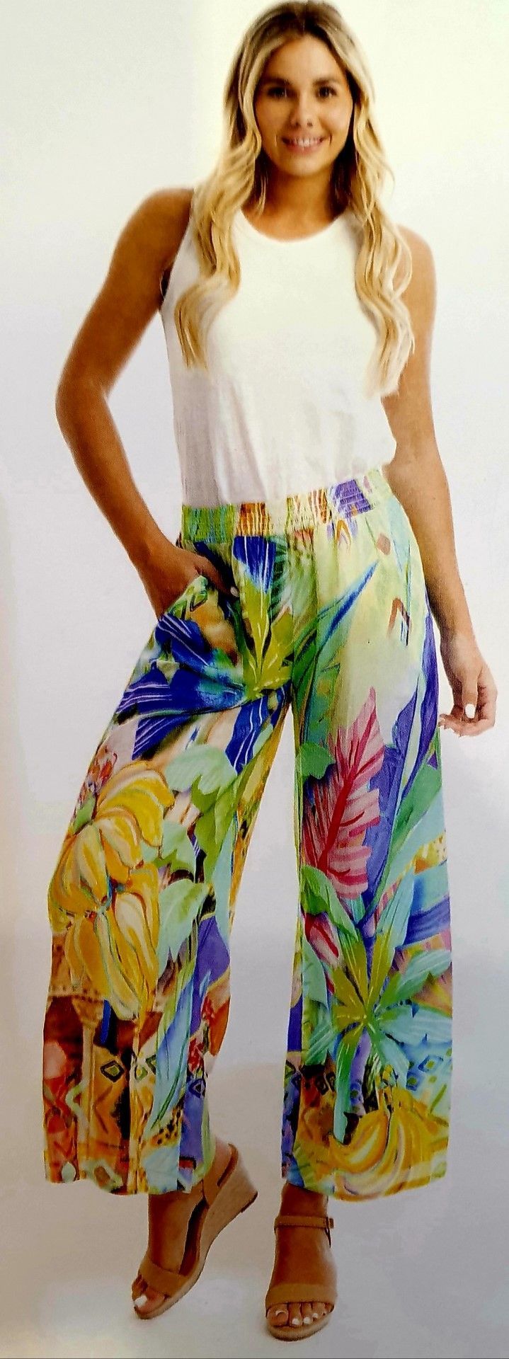 Woman in white top and colorful floral pants, smiles, stands indoors.