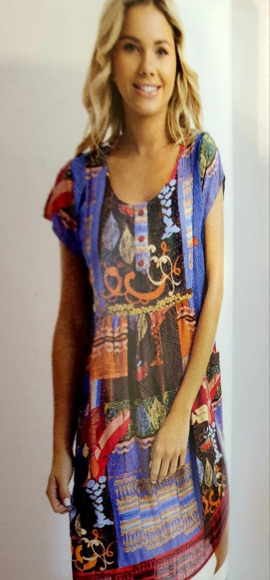 Blonde woman wearing a colorful patterned tunic dress, standing against a white wall.