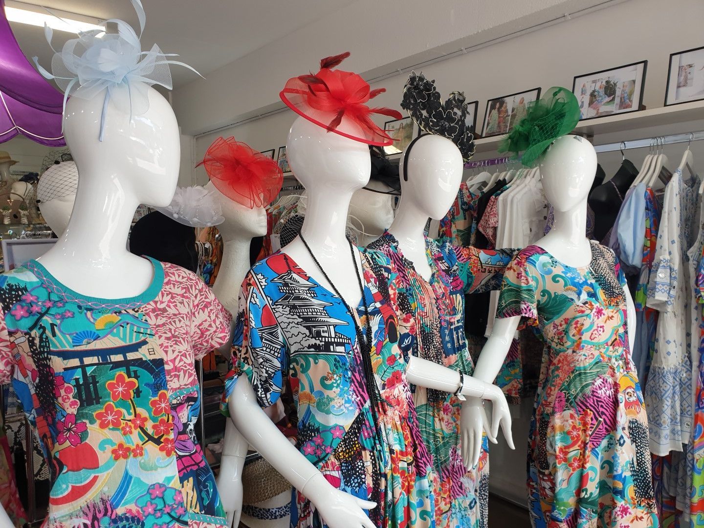 Mannequins in a shop wearing colorful dresses and hats; clothes rack in the background.