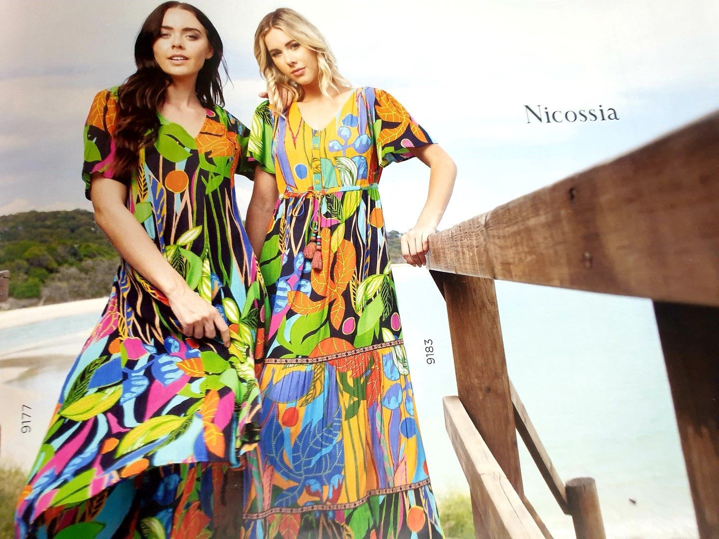 Two women in vibrant floral dresses stand on a wooden deck overlooking the ocean.