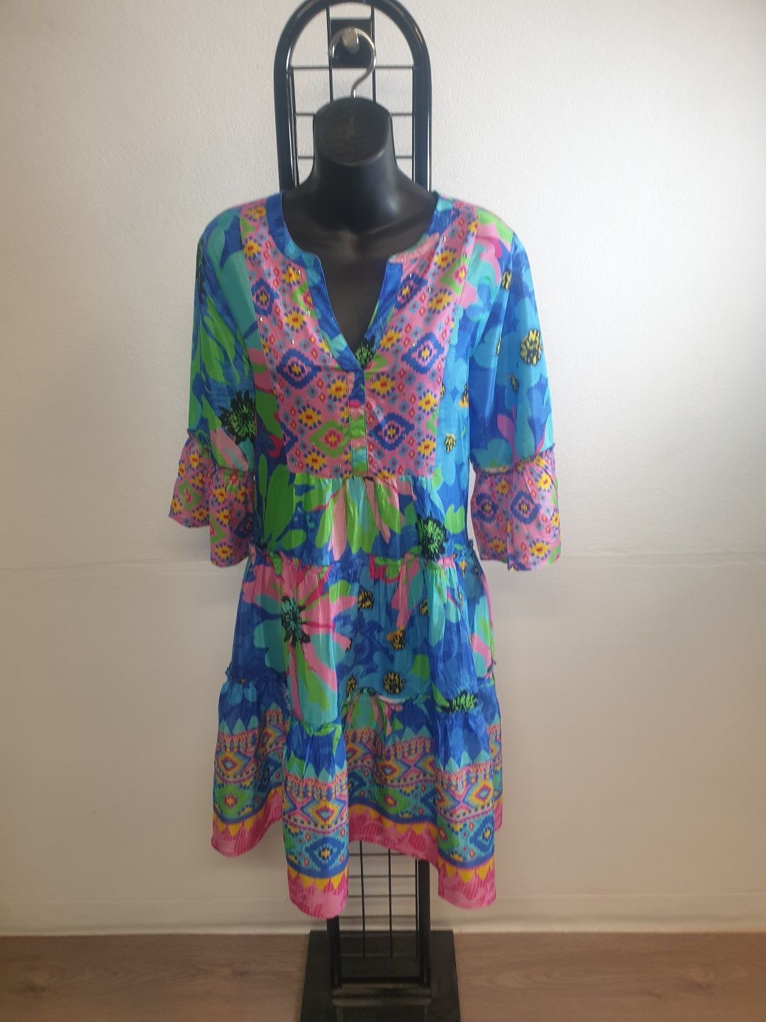Blue, pink, and green patterned dress on a black mannequin, on a black display stand, against a white wall.