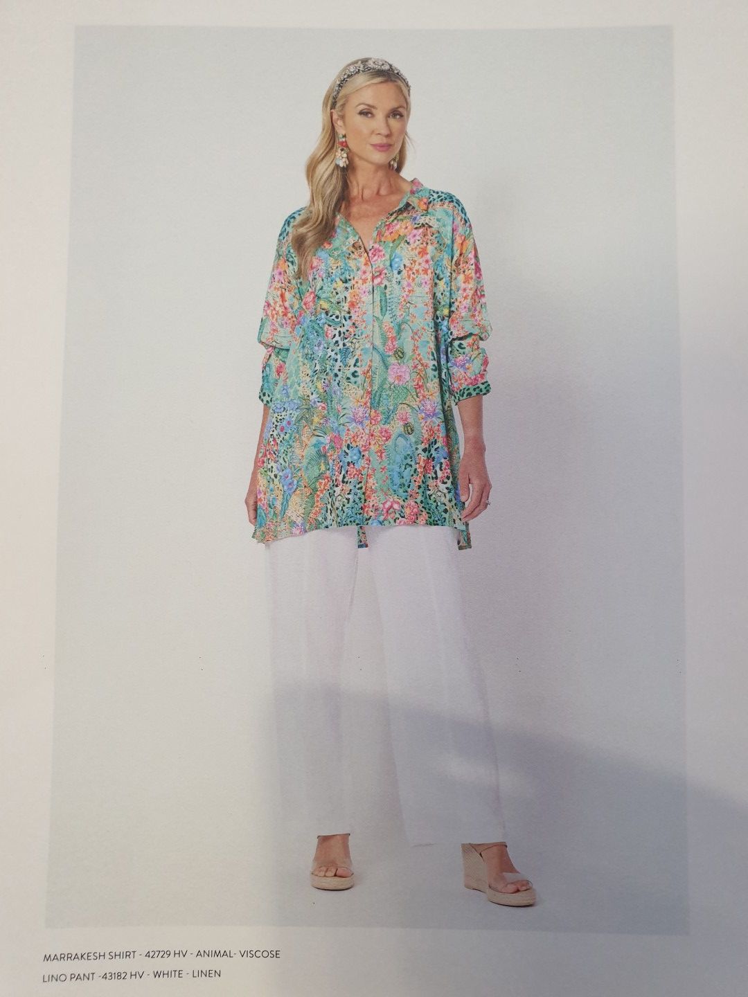 Woman in floral top and white pants stands, smiling. Light background.