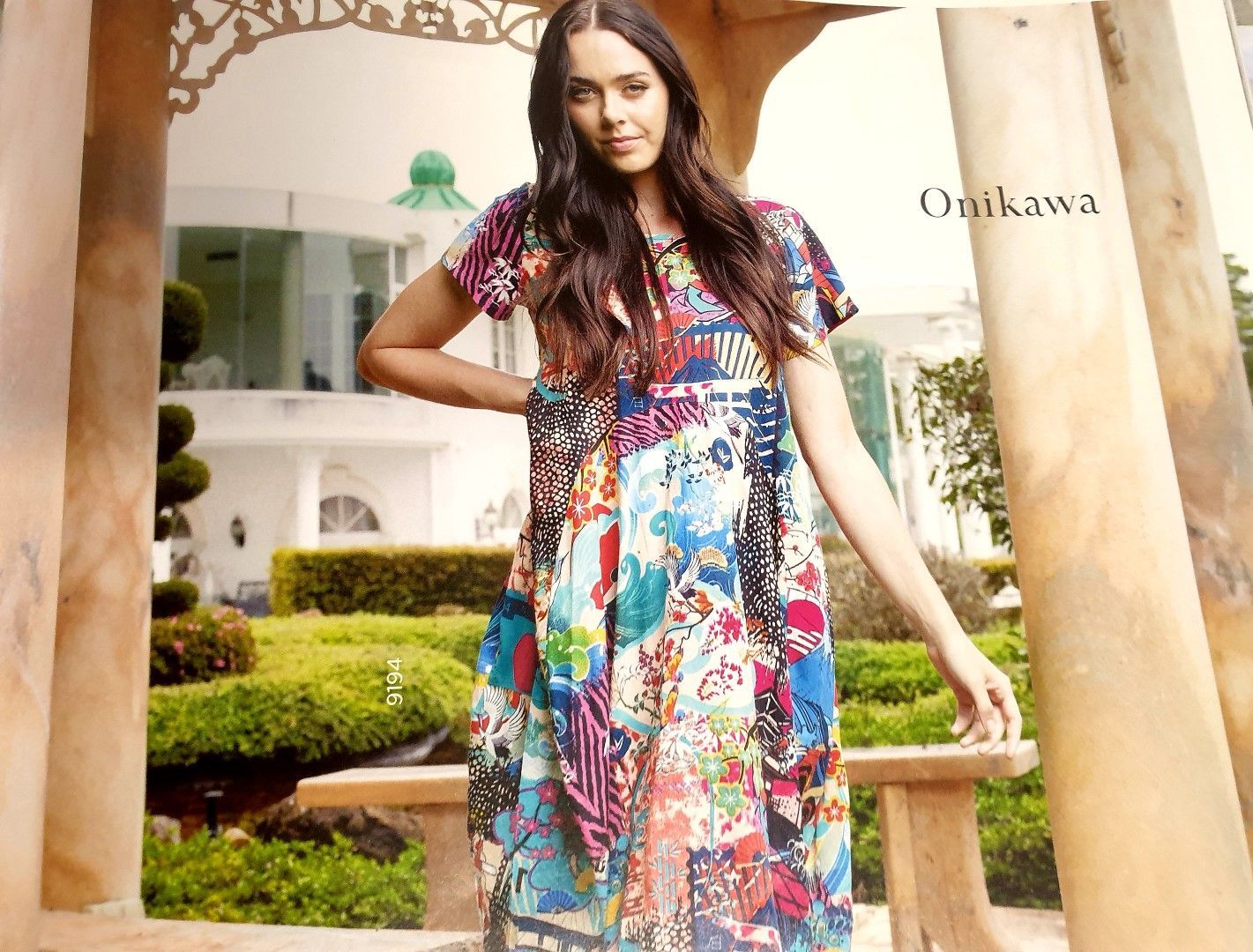 Woman in patchwork dress poses outdoors near a white house and garden.
