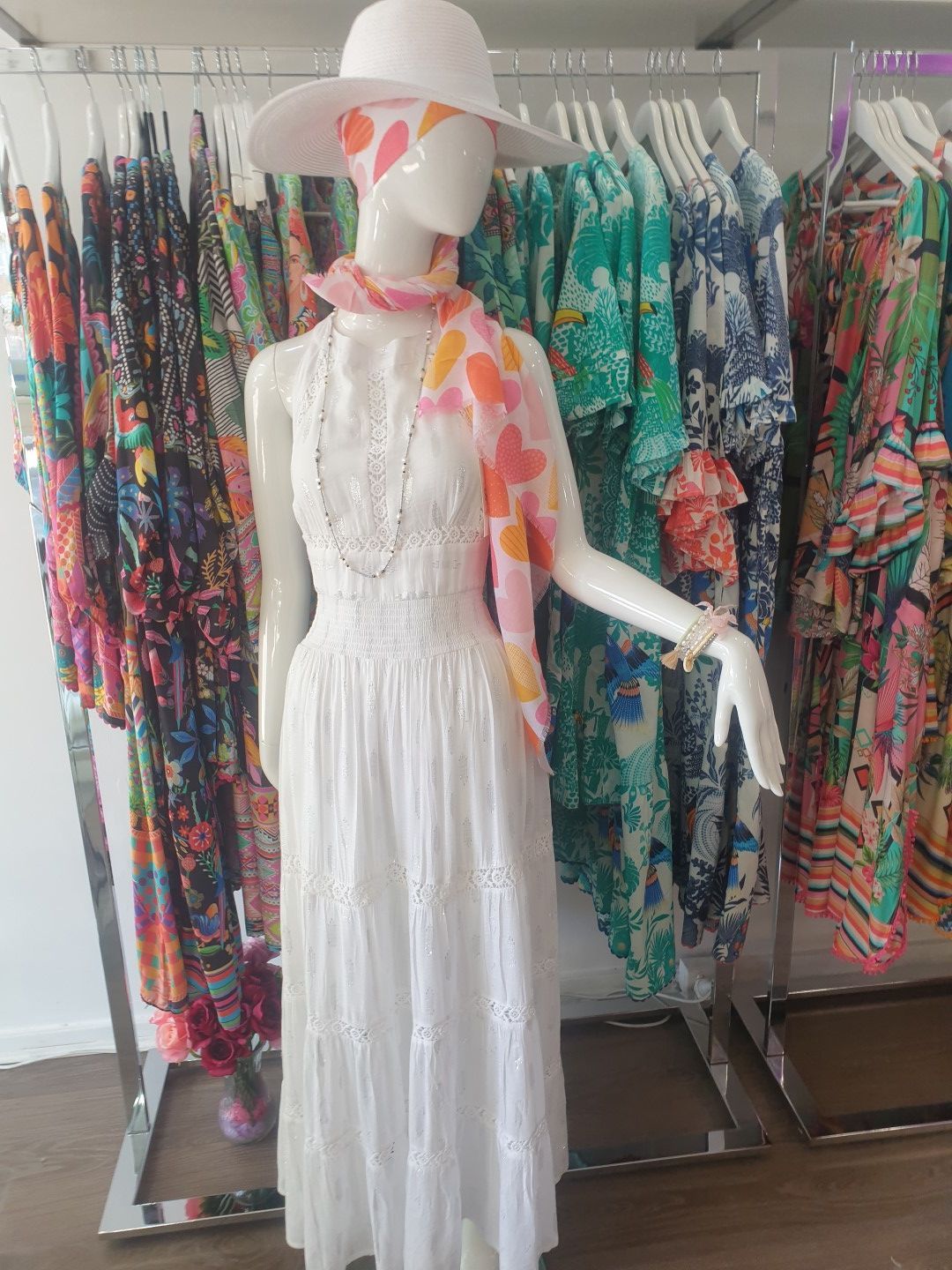 Mannequin in white dress, hat, and scarf, in a clothing store with colorful dresses on display.