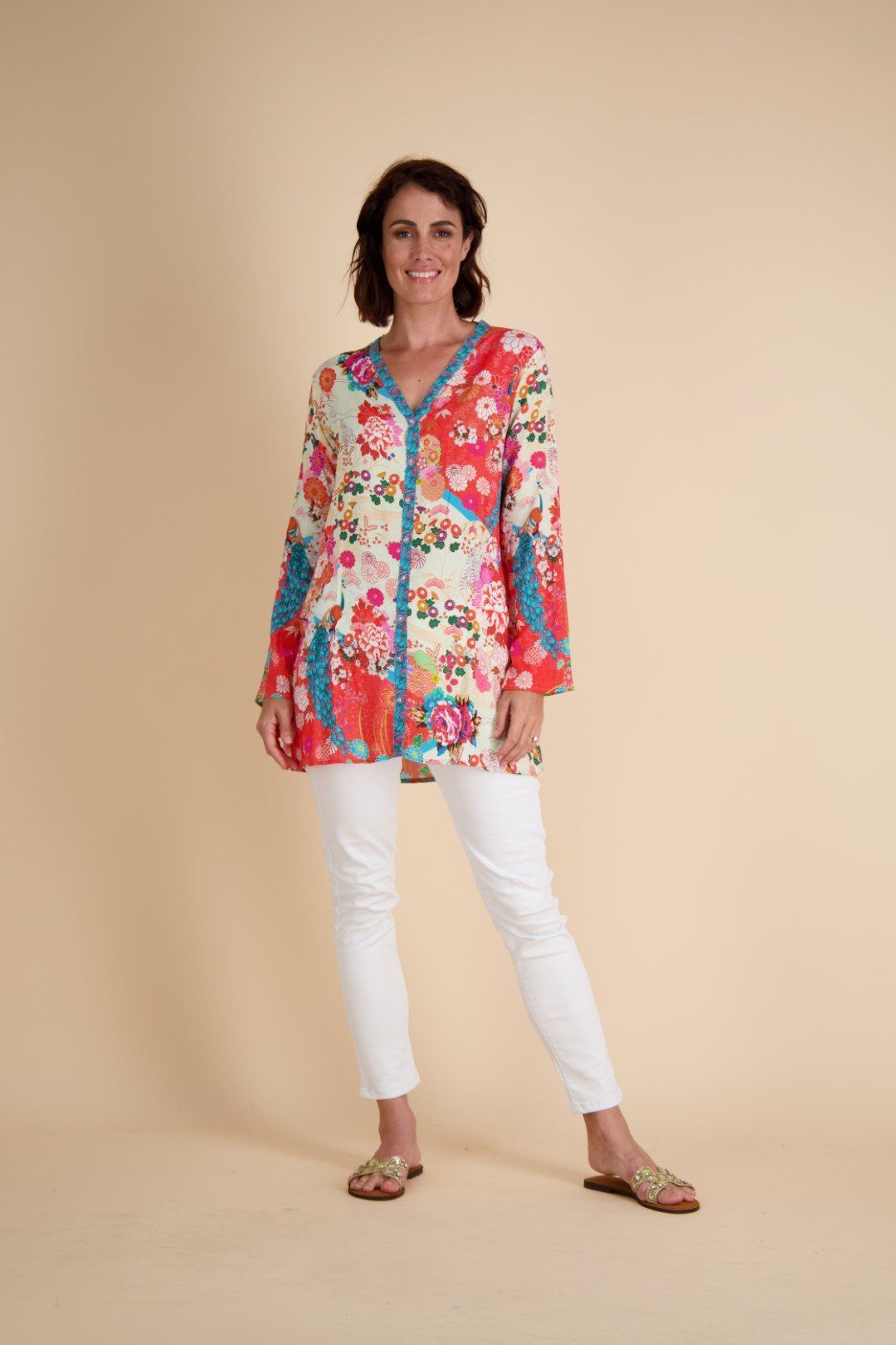 Woman in floral tunic and white pants, smiling, standing. Beige backdrop.