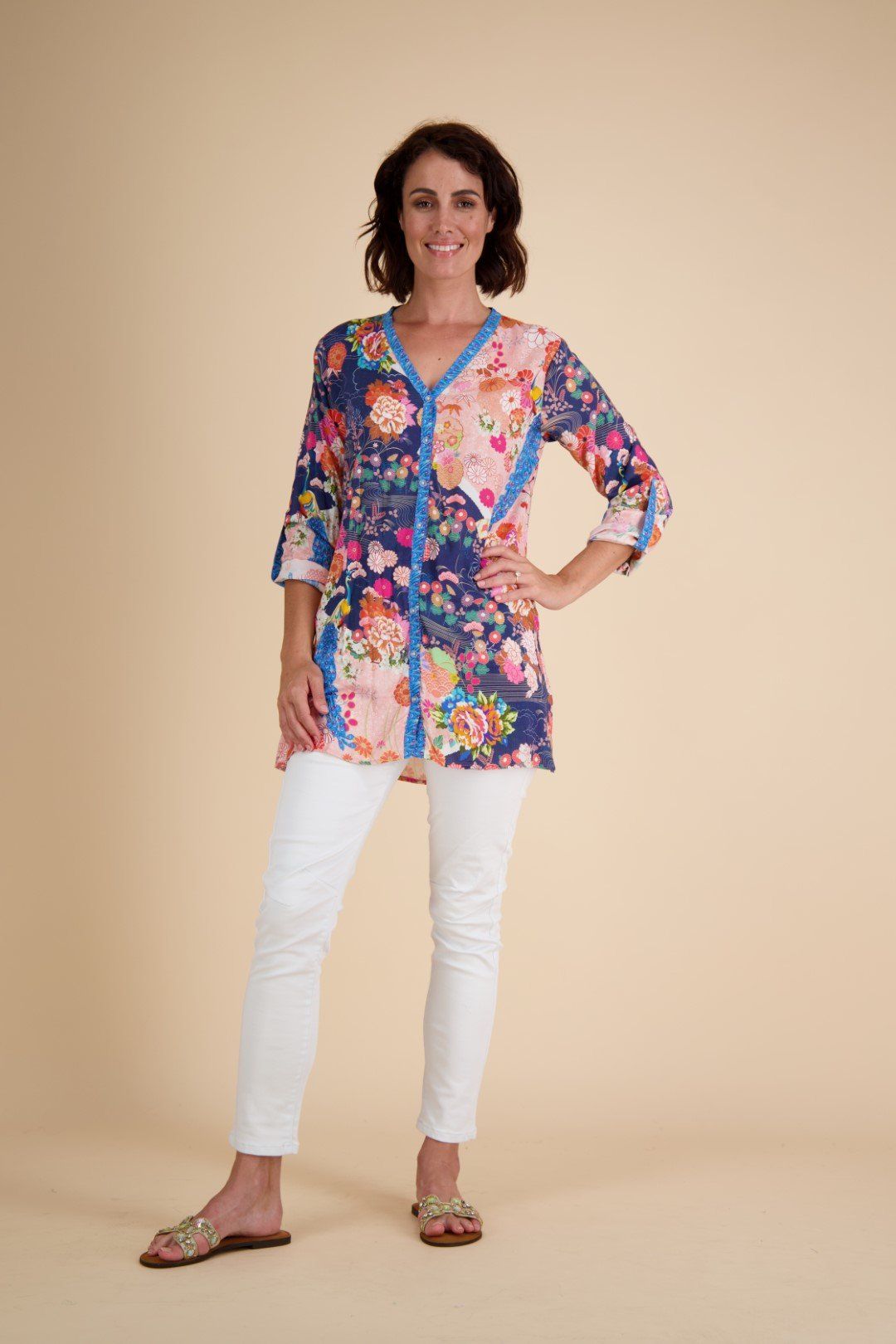 Woman in floral tunic and white pants, posing with hand on hip. Beige background.