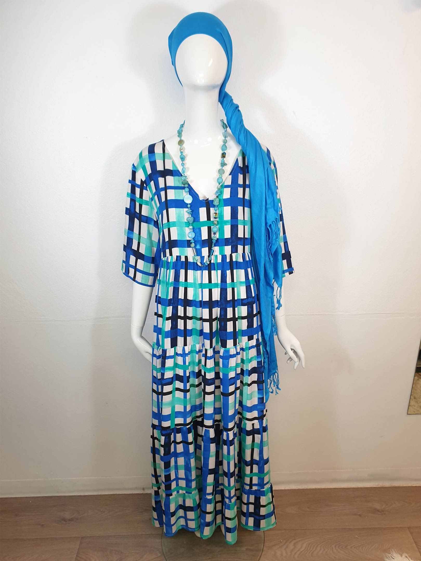 Mannequin wearing a long blue and white plaid dress, turquoise headscarf, and beaded necklace.
