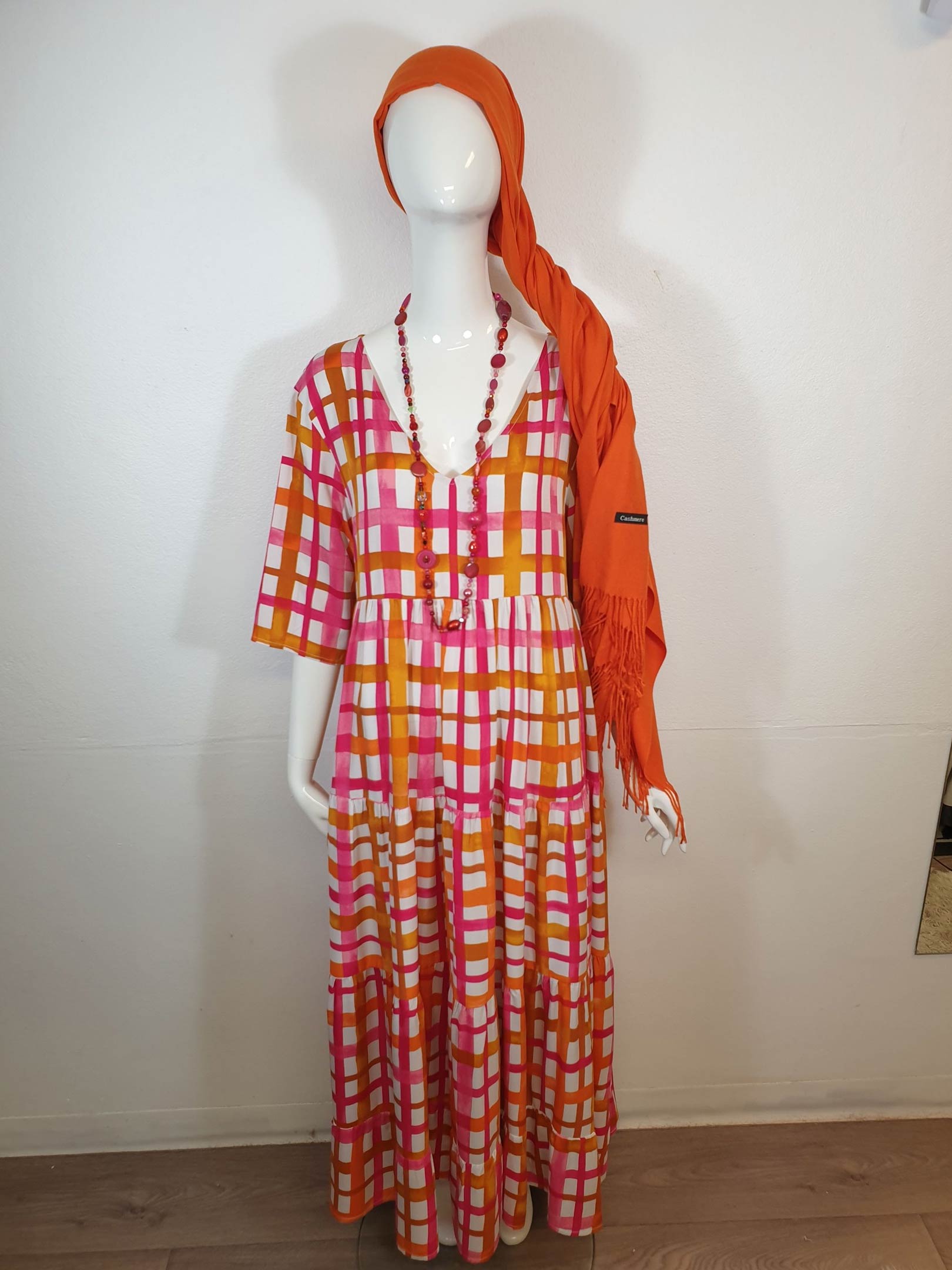 Mannequin in a colorful plaid dress, orange head wrap, and beaded necklace against a white wall.