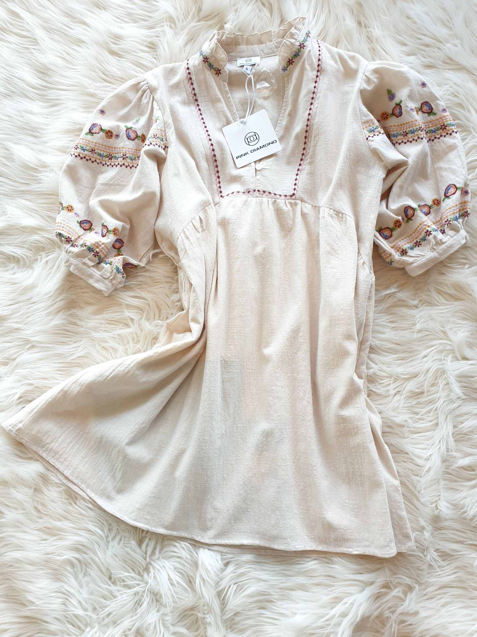 Cream-colored dress with embroidered floral sleeves and neckline. Lying on a fluffy, white surface.