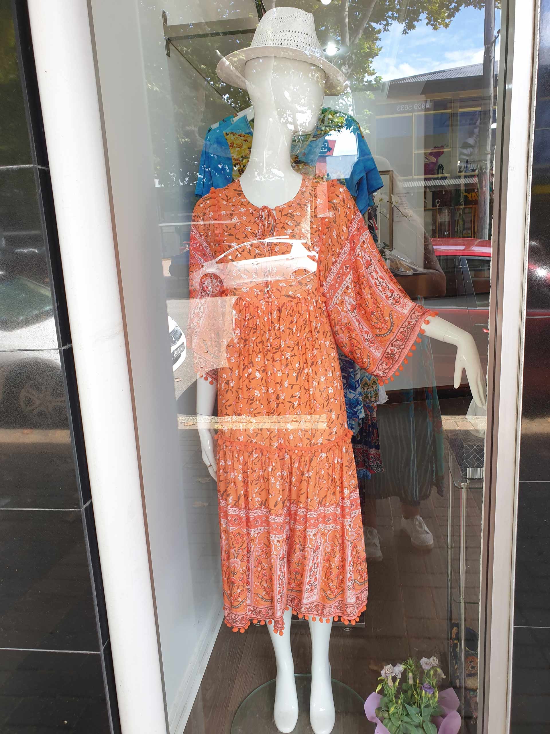 Mannequin in an orange patterned dress and white hat inside a storefront window.
