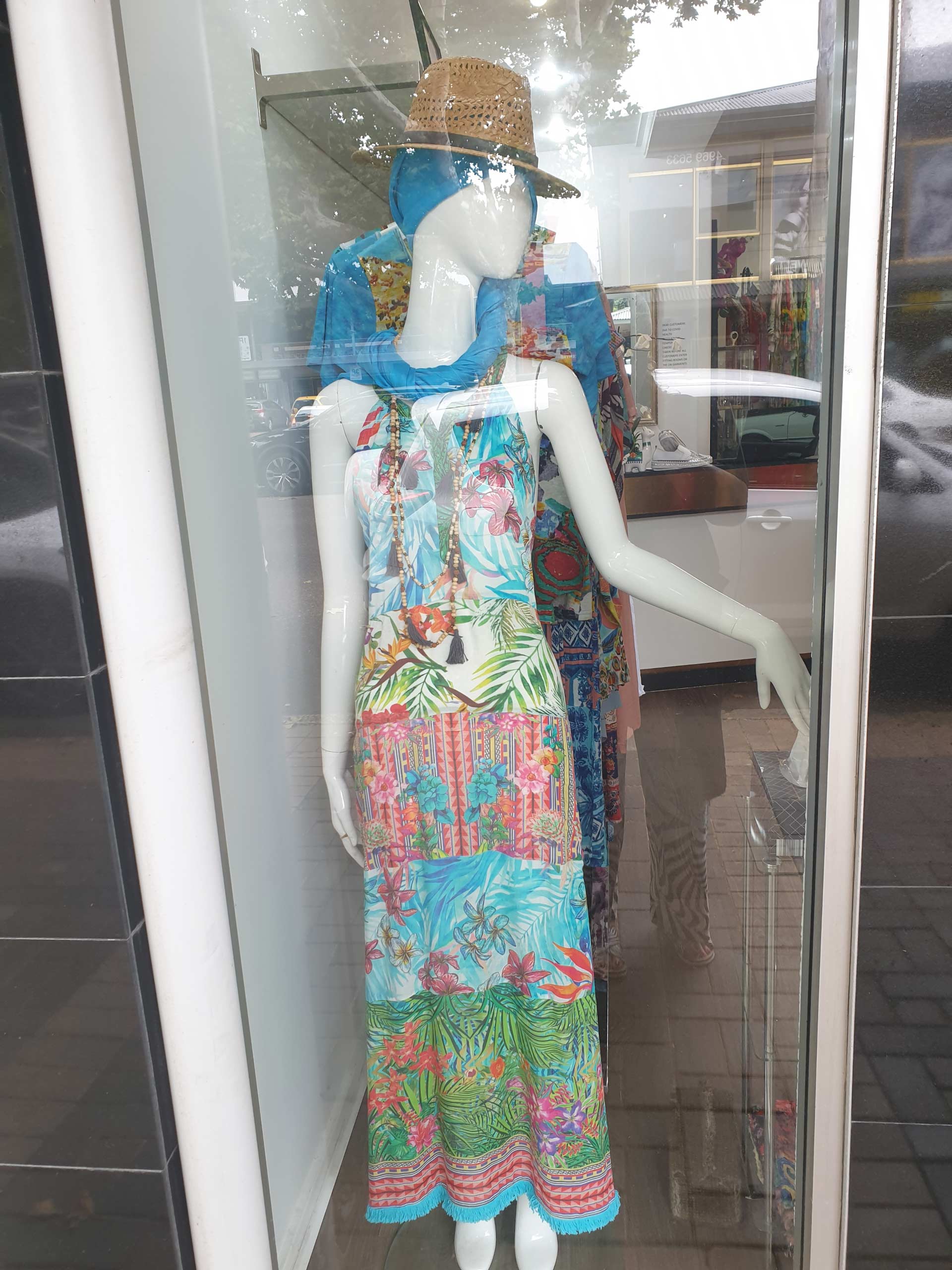 Mannequin in a window wearing a colorful patterned maxi dress and blue scarf, with a hat.