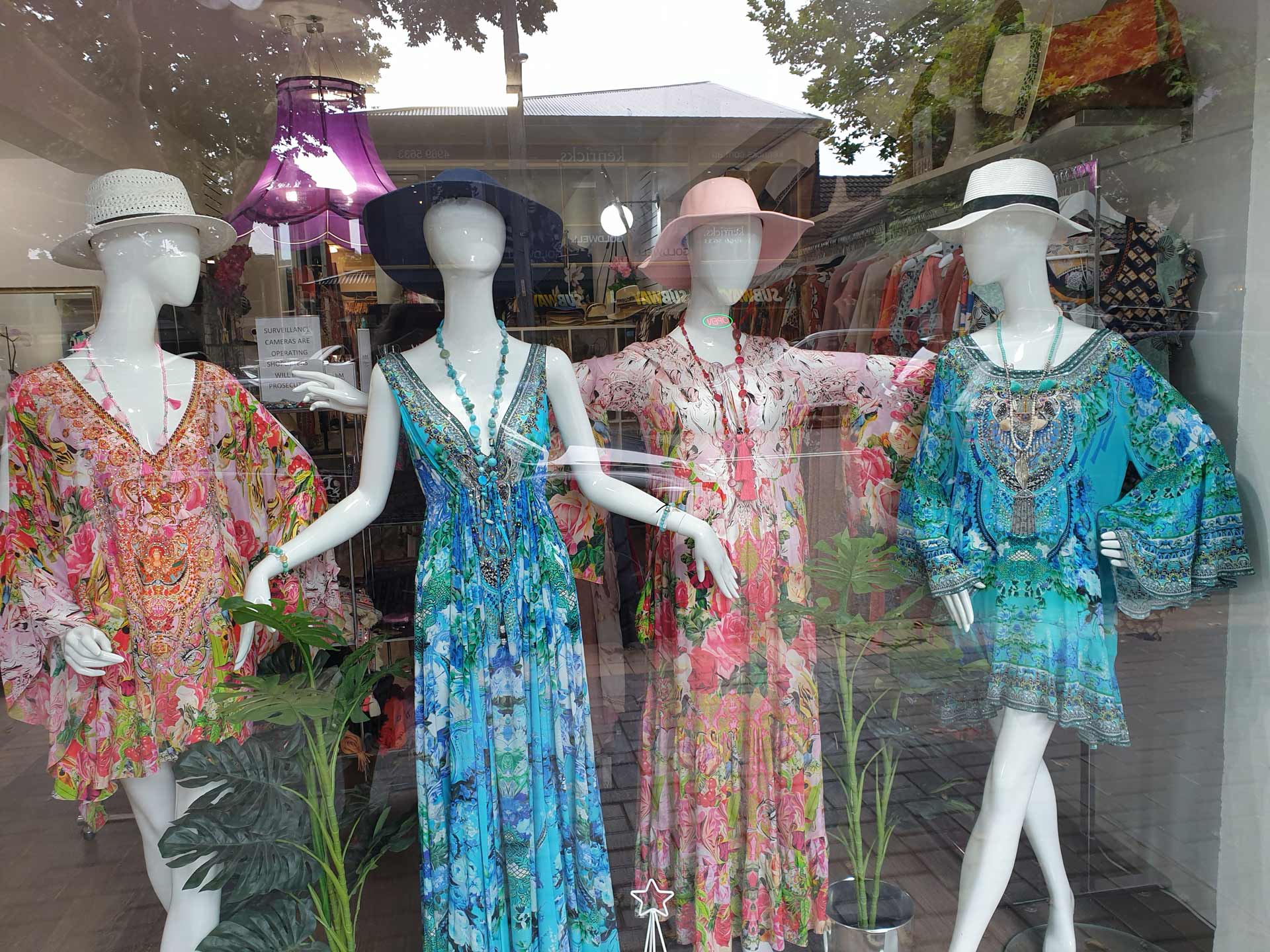Four mannequins in a shop window wearing colorful, patterned dresses and hats.