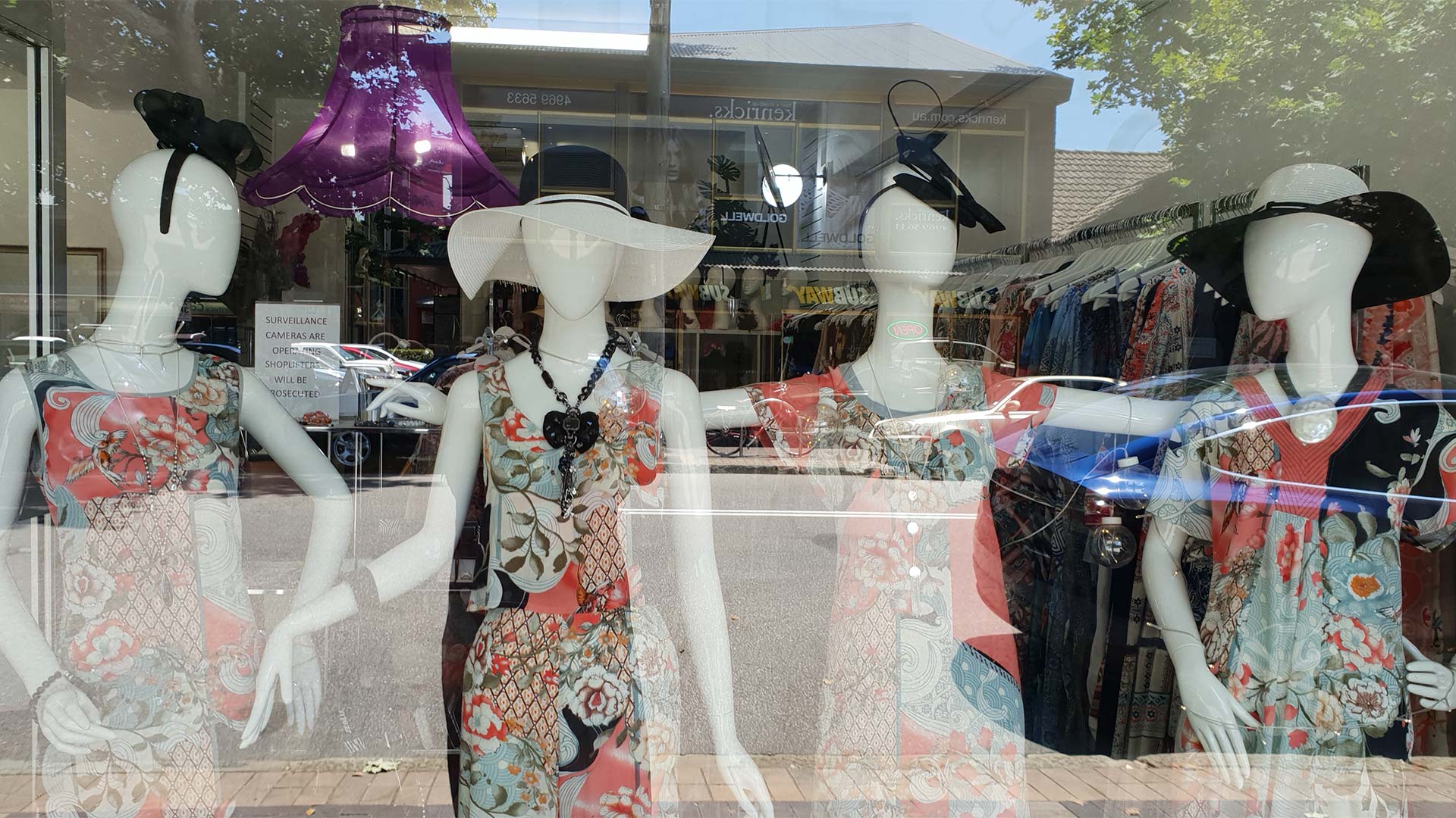 Storefront window display with mannequins wearing floral dresses and hats.