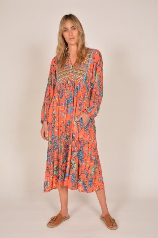 Woman in orange floral dress, standing indoors, smiling, wearing sandals.