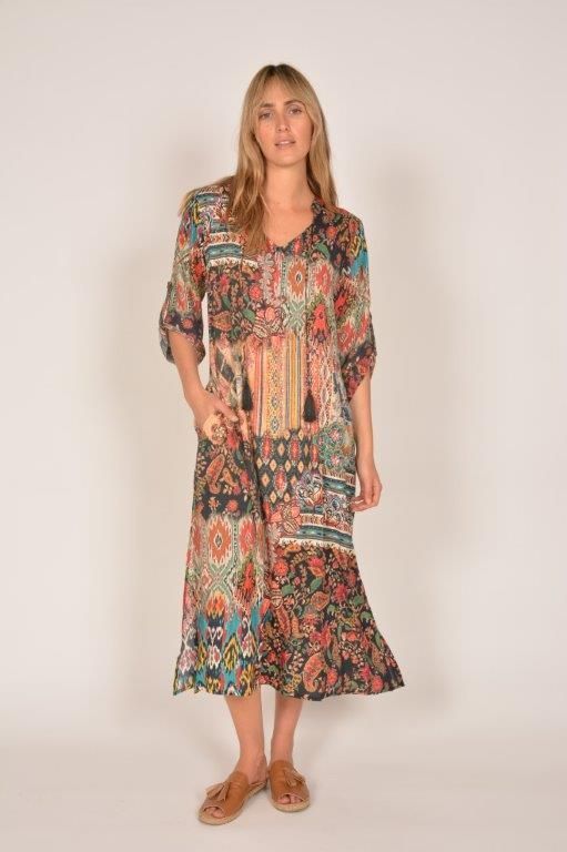 Woman wearing a colorful, patterned dress with tassels and sandals, standing indoors.