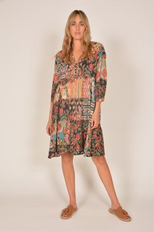 Woman in a colorful patterned dress stands against a white backdrop, wearing sandals.