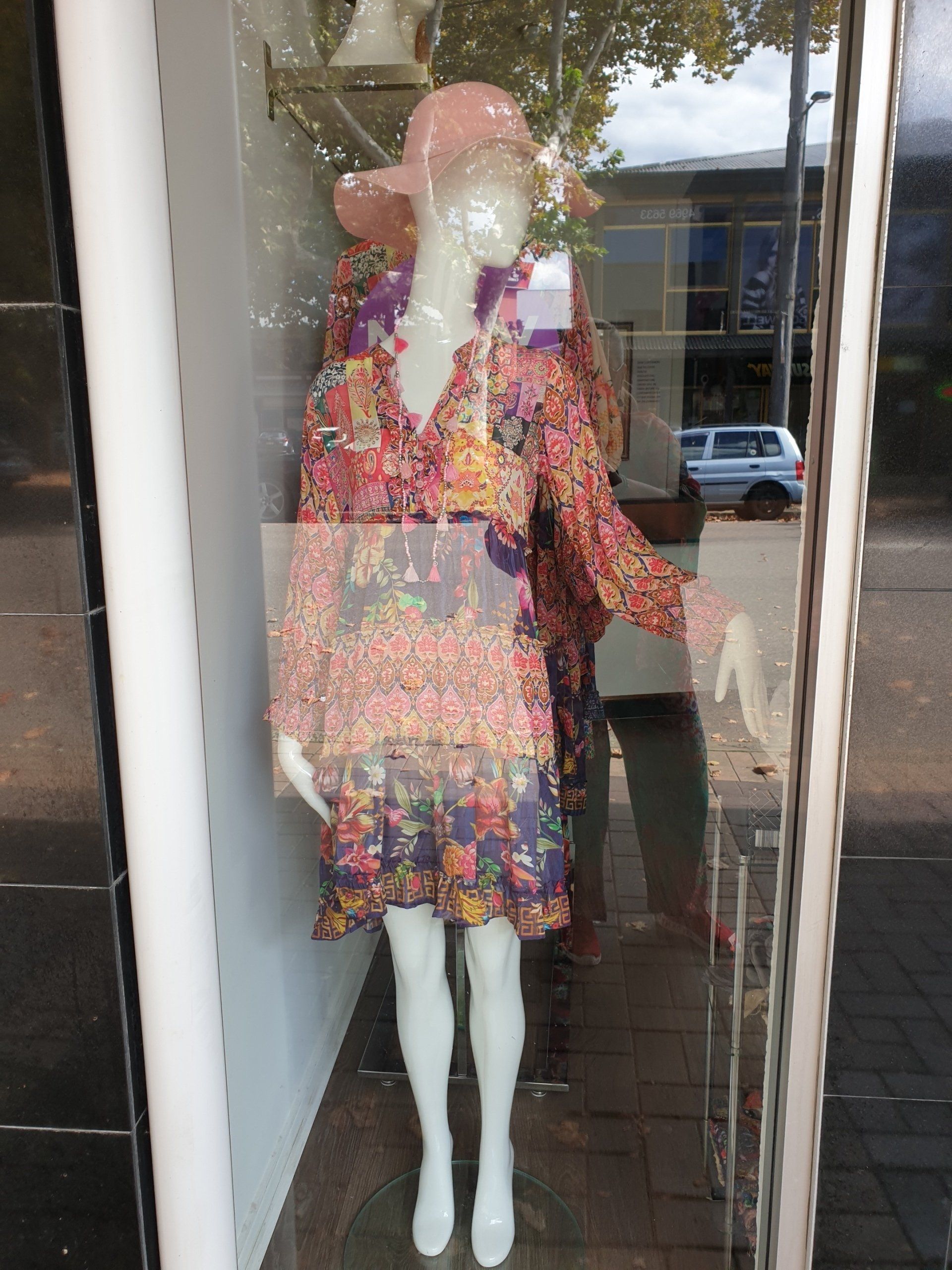 Mannequin in floral dress and pink hat in a shop window, with a reflected street view.