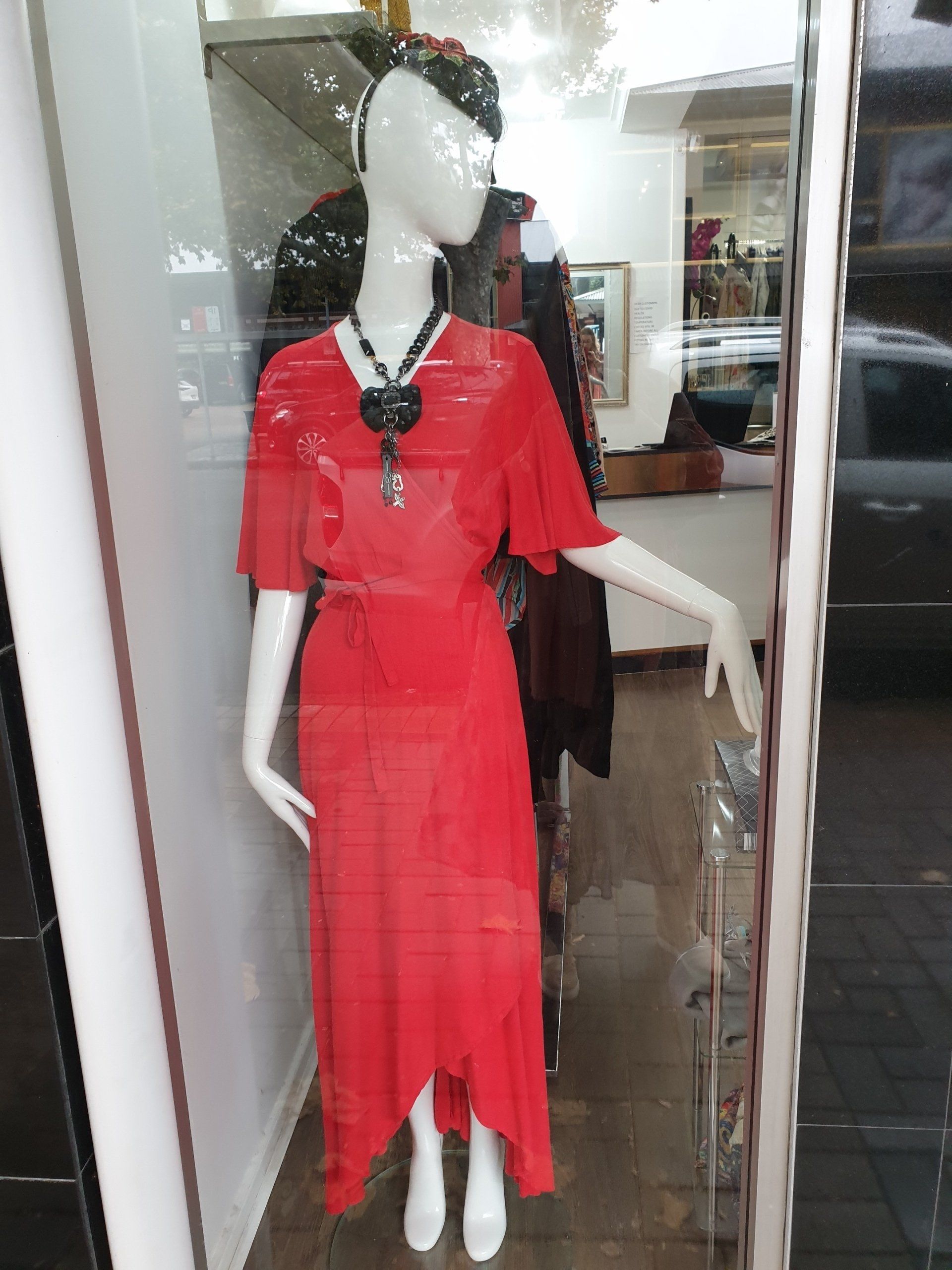 A red dress on a mannequin in a shop window. The mannequin wears a hat and necklace.