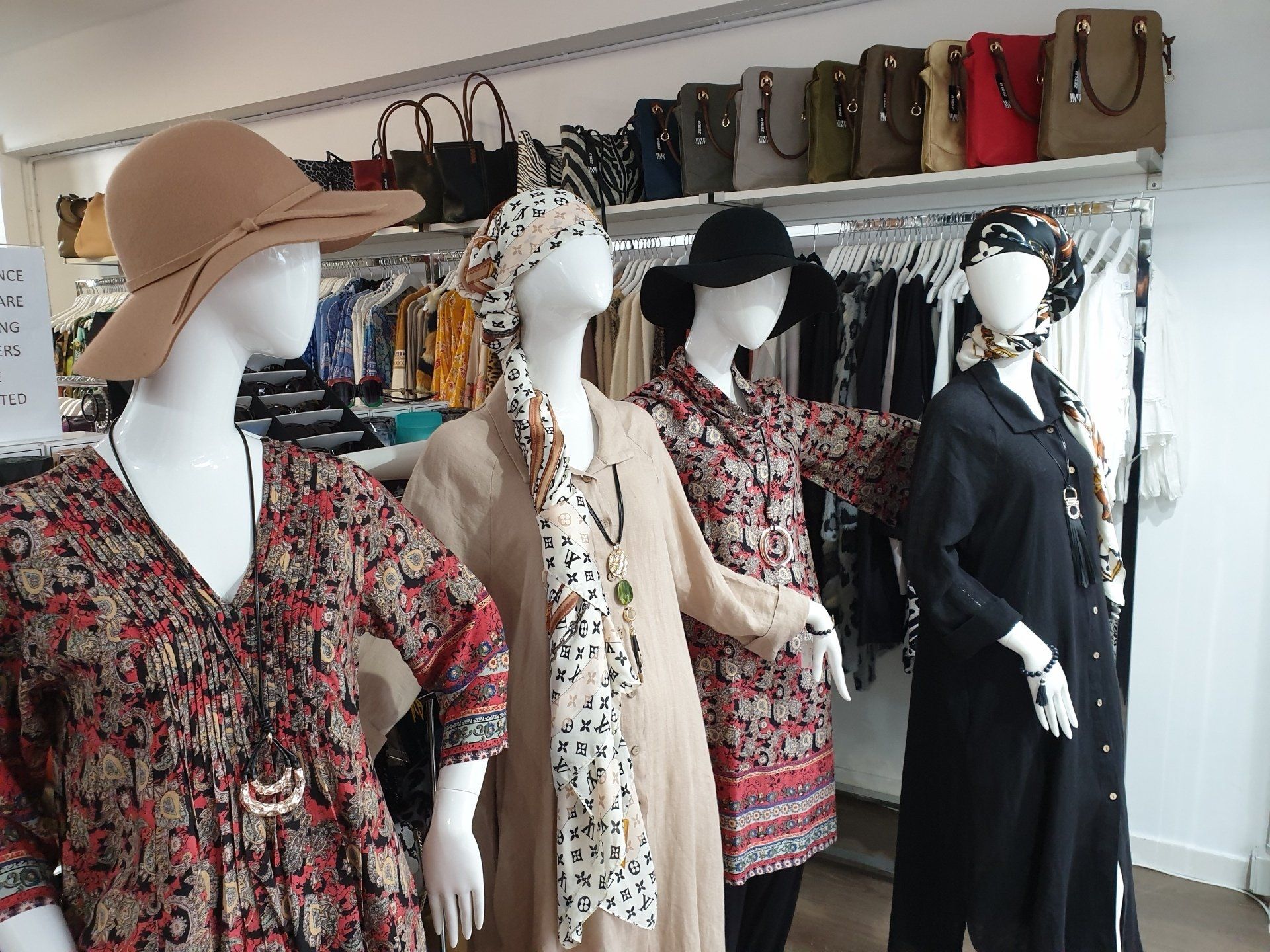 Four mannequins in a clothing store window; wearing outfits, hats, and scarves; merchandise on racks and shelves.