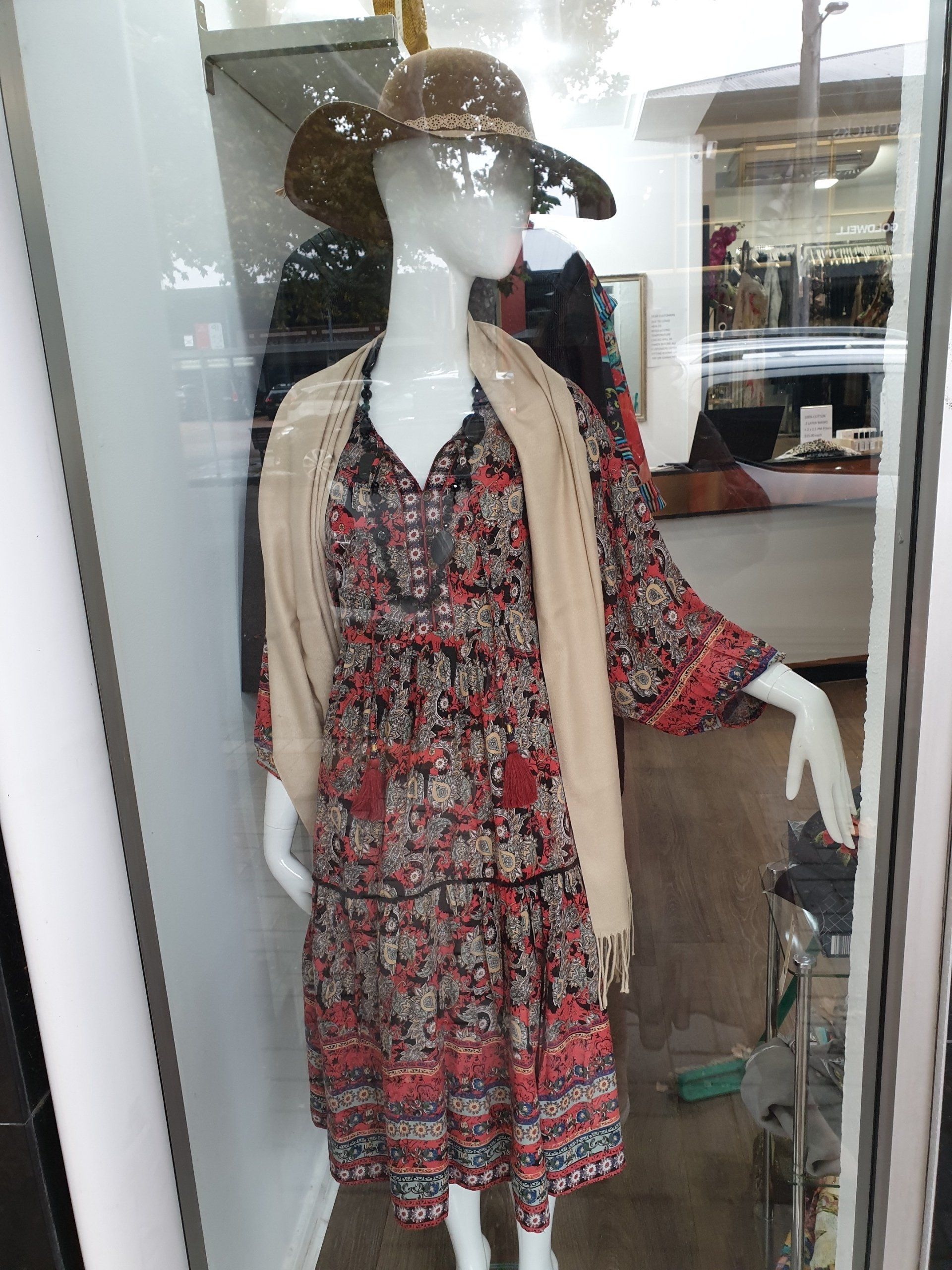 Mannequin in a shop window wearing a patterned dress, a tan shawl, and a straw hat.