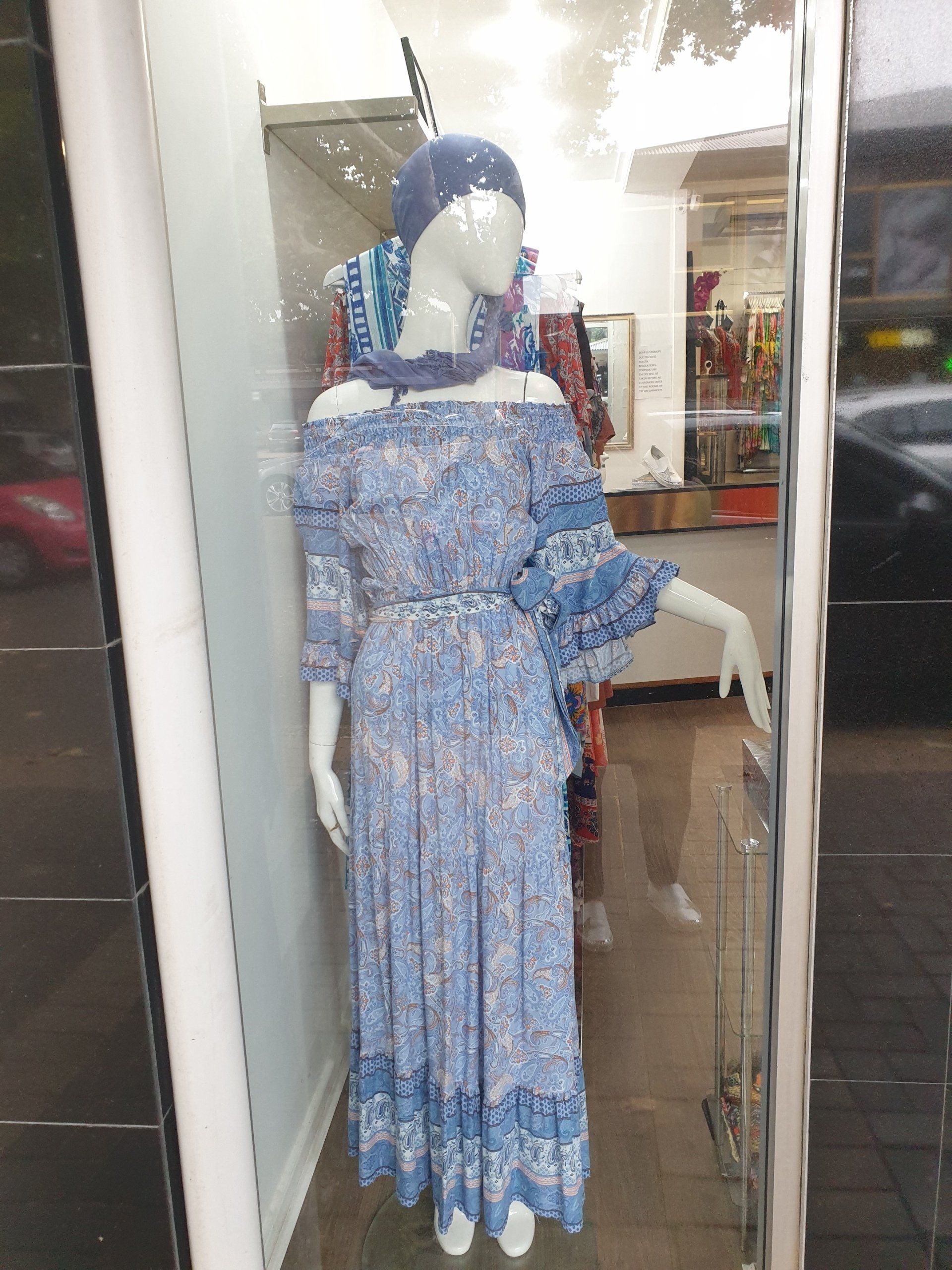 Mannequin in blue floral off-the-shoulder dress, hat, and jewelry in a shop window.