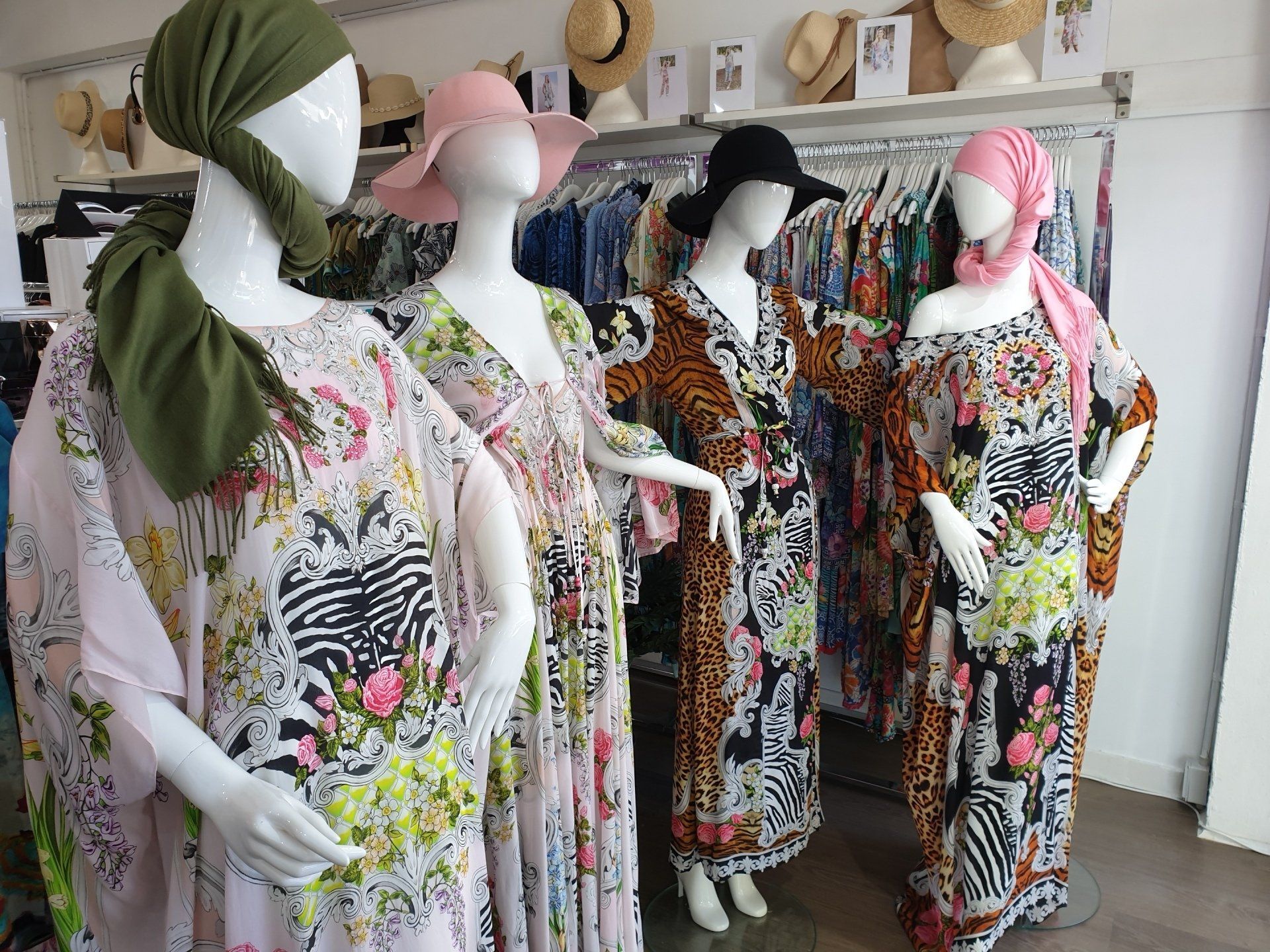 Four mannequins wearing colorful, patterned kaftans in a clothing store; various hats hang in the background.