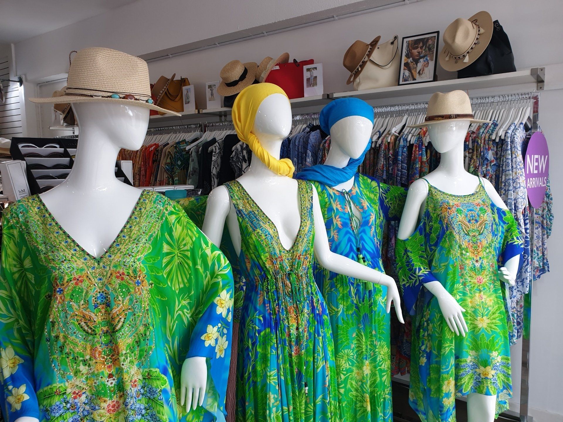 Four mannequins in a boutique, wearing vibrant green and blue dresses, straw hats.