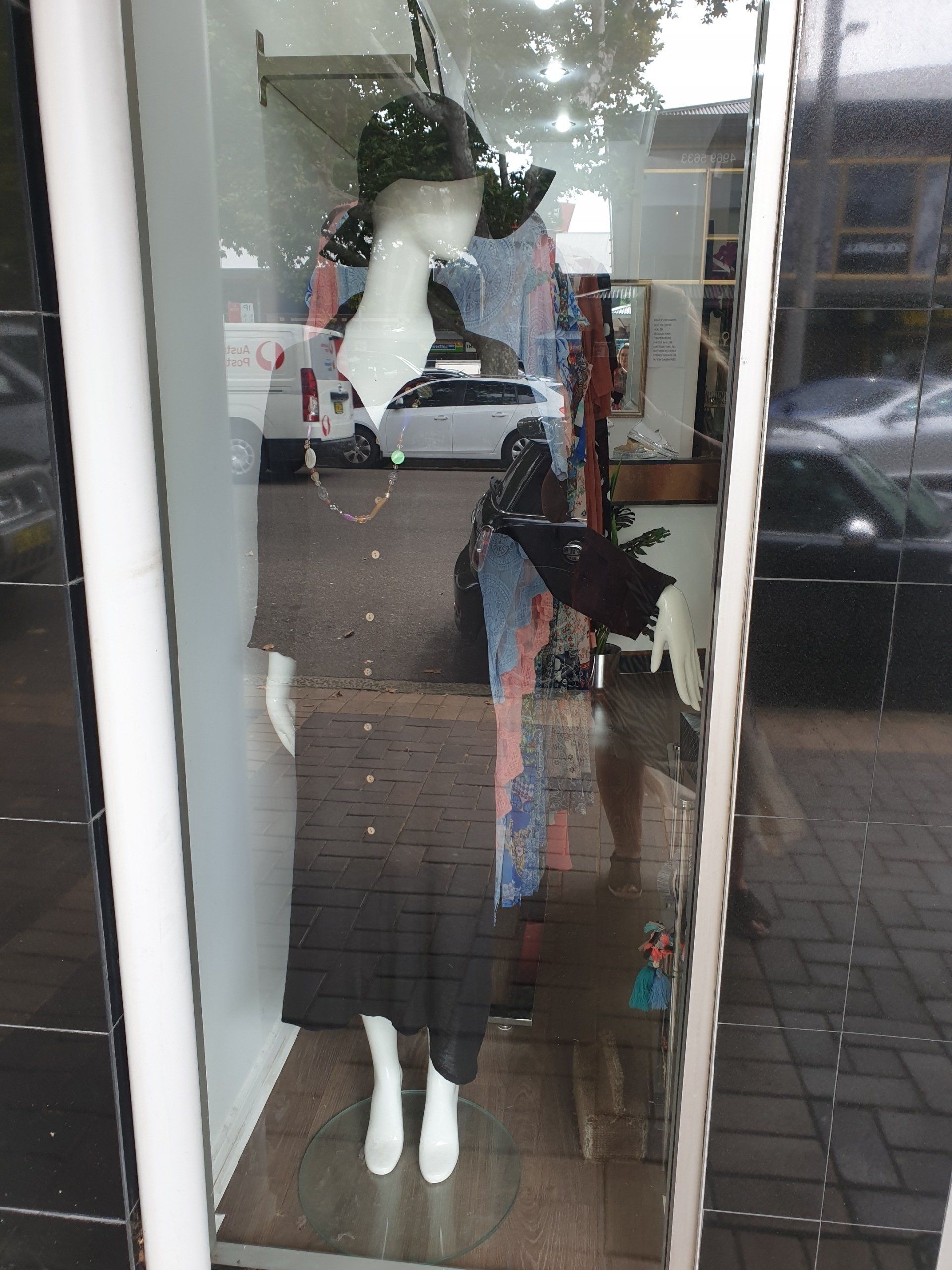 Mannequin in shop window wearing a dark dress and hat. Street reflection visible.
