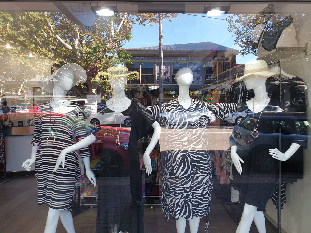 Mannequins in a shop window displaying summer dresses, sunhats, and jewelry; outdoors in a sunny setting.