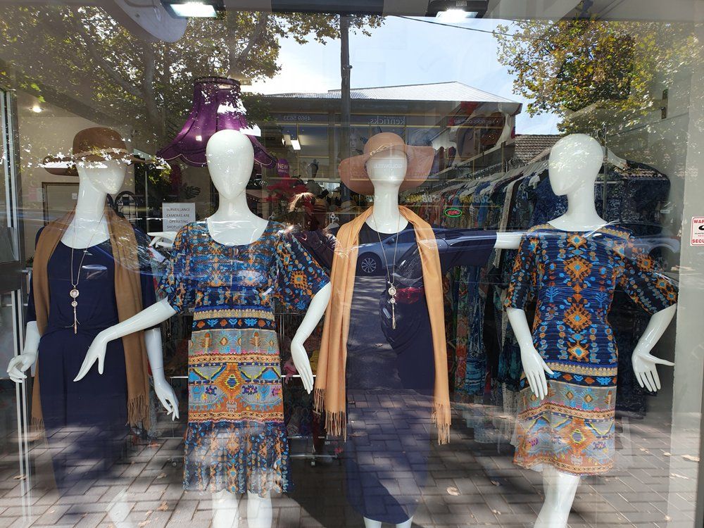 Four mannequins in a shop window display dresses, shawls, and hats. Trees and buildings reflect in the glass.