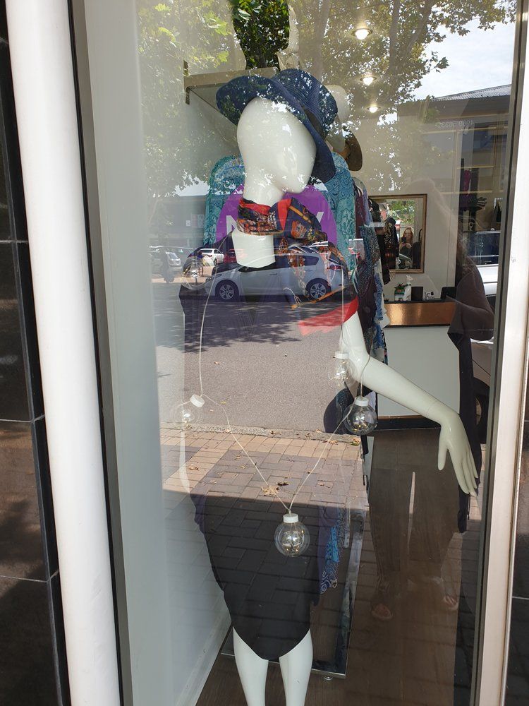 Mannequin in a shop window, wearing a colorful top, hat, and black skirt; street scene reflected.