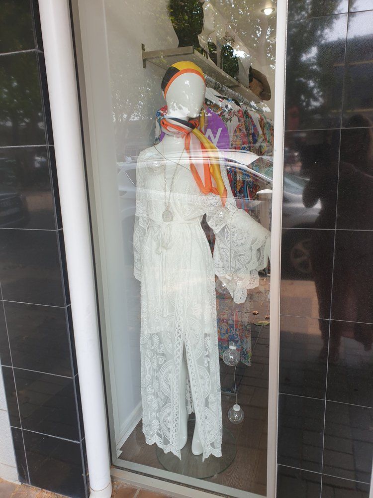 Mannequin in white lace dress, window display. Headscarf, scarf, clothes rack visible.