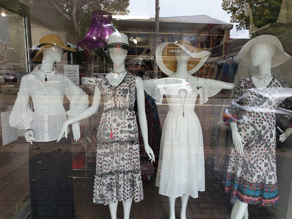 Four mannequins in a shop window display dresses and hats; white, floral, and neutral colors.